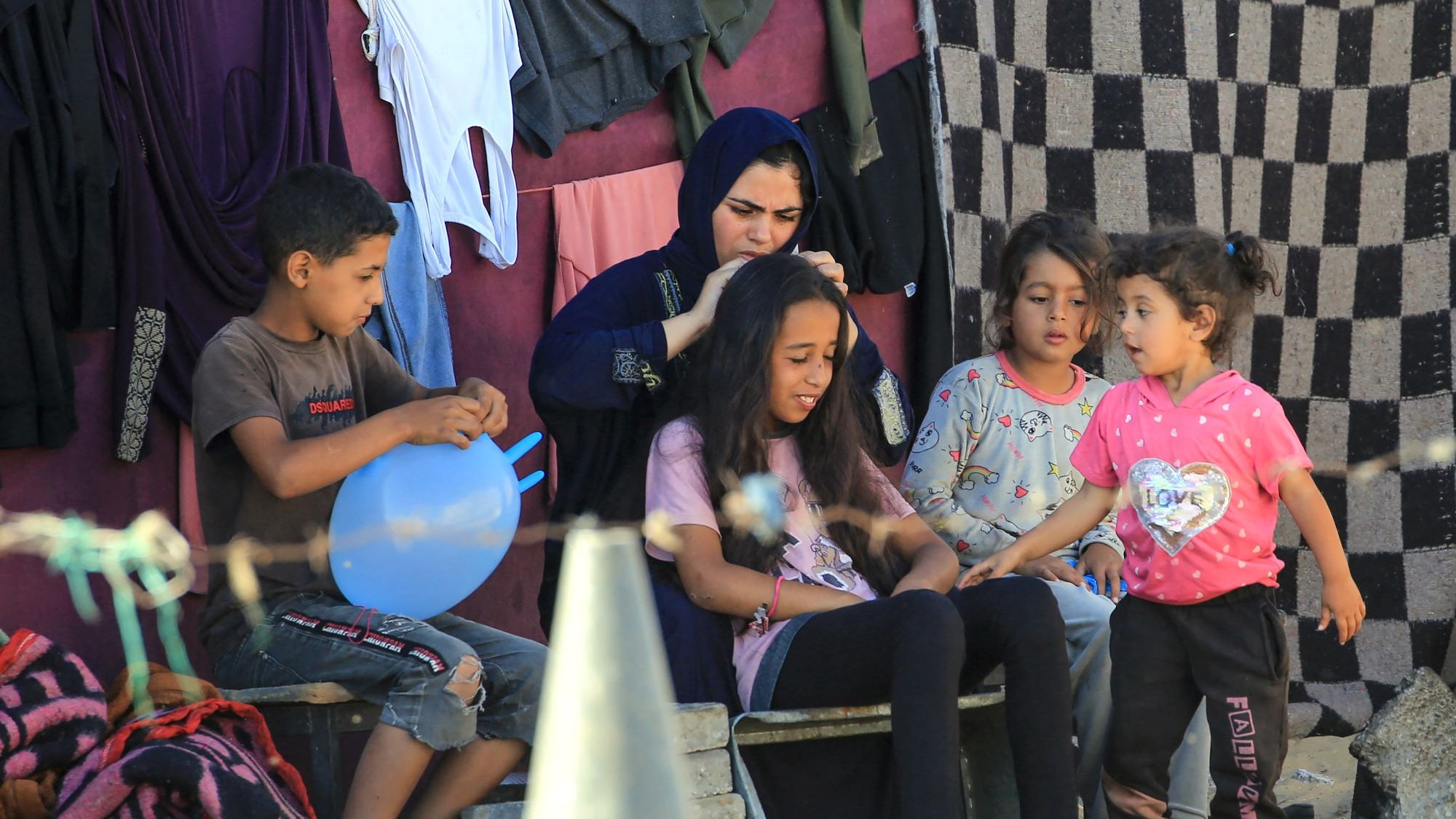 L'image montre un groupe d'enfants et une femme assis à l'extérieur, dans un environnement simple. Les enfants semblent interagir joyeusement, avec une petite fille qui parle à une autre. Une femme en vêtements traditionnels s'occupe des cheveux d'une jeune fille assise devant elle. En arrière-plan, on peut voir des vêtements accrochés et un sol en sable. L'ambiance est conviviale et émotive, évoquant des moments de partage et de complicité.