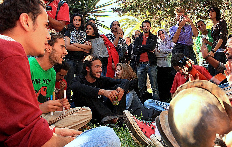 L'image montre un groupe de personnes rassemblées dans un environnement extérieur, probablement dans un parc ou un jardin. Des jeunes, assis sur l'herbe, semblent participer à une activité musicale, avec des instruments comme une guitare et des percussions. Certains d'entre eux sont engagés dans une discussion animée. En arrière-plan, d'autres personnes regardent ou écoutent, créant une atmosphère conviviale et collective. Les expressions des participants varient, allant de la concentration à l'amusement.