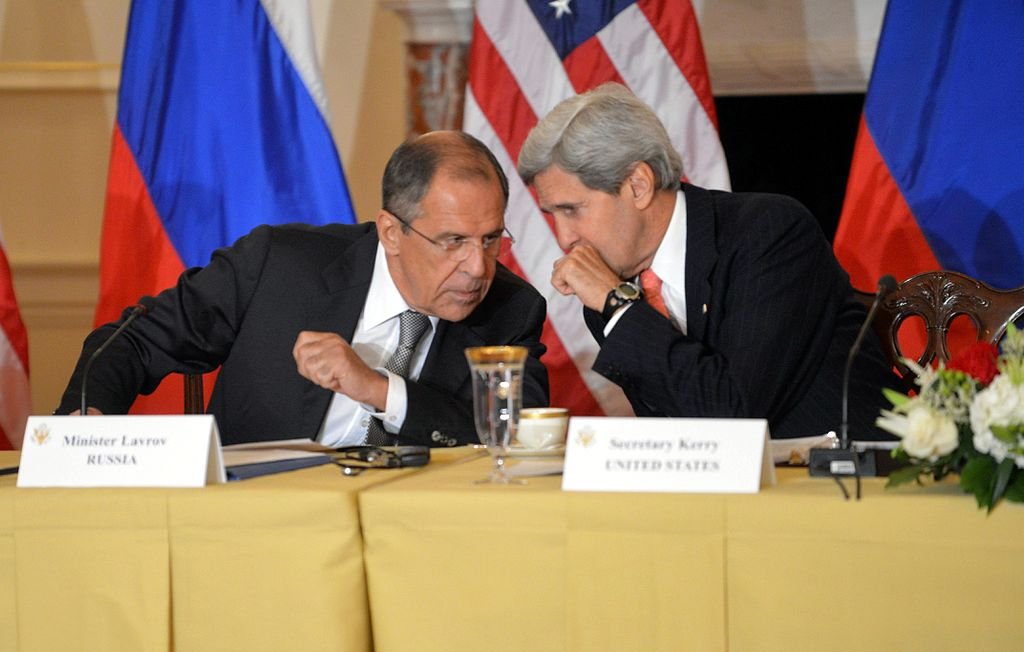 L'image montre deux hommes assis à une table, engageant une conversation. L'un est identifié comme étant le ministre des Affaires étrangères de la Russie, Sergueï Lavrov, et l'autre comme John Kerry, l'ancien secrétaire d'État américain. Ils semblent discuter de manière confidentielle, entourés de décorations représentant les drapeaux de la Russie et des États-Unis. Sur la table, il y a des documents et une tasse. L'atmosphère semble sérieuse et diplomatique.