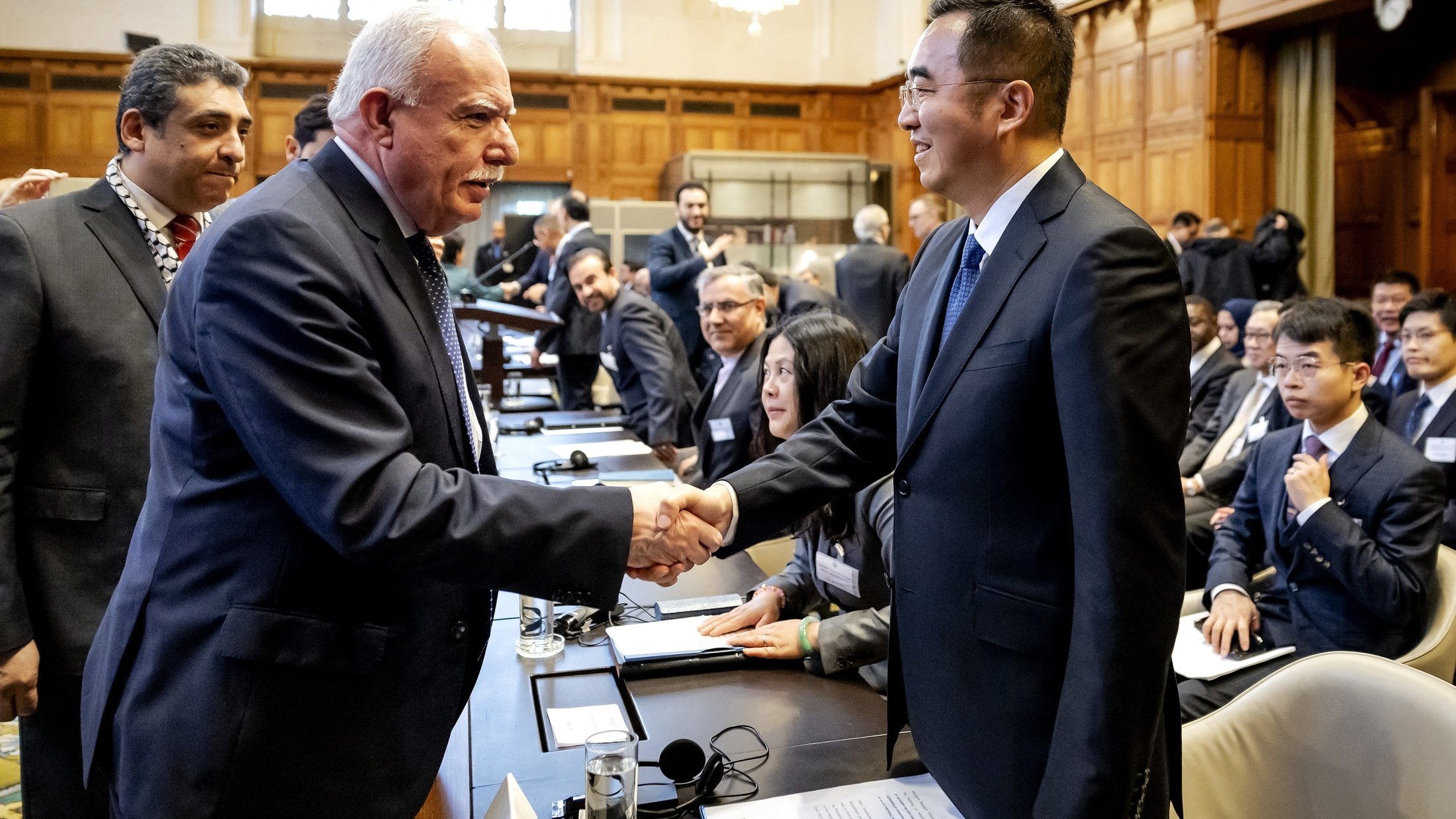 L'image montre une scène de réunion ou de conférence, probablement dans un cadre formel. Deux hommes se serrent la main, souriant, ce qui évoque une interaction chaleureuse ou un accord. En arrière-plan, on aperçoit d'autres participants qui semblent également engagés dans des discussions. La salle est spacieuse, avec une décoration élégante, des fenêtres laissant passer une lumière naturelle, et des sièges disposés autour d'une grande table. L'atmosphère semble professionnelle et collaborative.