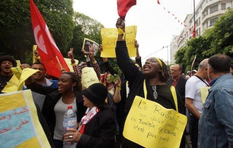 L'image montre une manifestation dans une rue animée, avec des gens qui tiennent des pancartes et des drapeaux. Une personne au centre, habillée en noir, lève un panneau sur lequel est écrit "Le Racisme est un délit et un crime en Tunisie". Les manifestants semblent exprimer des préoccupations concernant le racisme. L'ambiance est vibrant et engagée, avec des participants qui chantent ou crient des slogans. Les éléments visuels suggèrent une mobilisation sociale pour des droits et la justice.