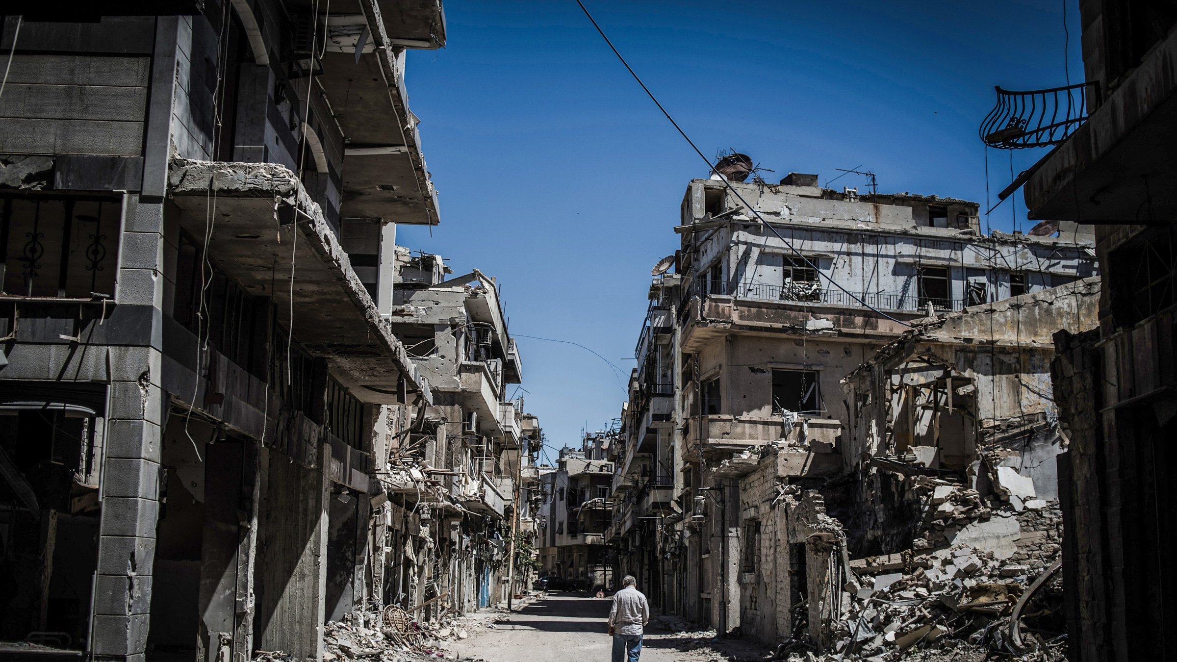 L'image montre une rue dévastée, bordée de bâtiments en ruine. Les murs sont fissurés et effondrés, témoignant des destructions causées par un conflit. Au centre de la scène, un homme marche seul, semblant contempler les vestiges de ce qui était autrefois un quartier vivant. Le ciel est clair, contrastant avec la désolation environnante. Cette image évoque une atmosphère de solitude et de tristesse face à la perte et à la destruction.