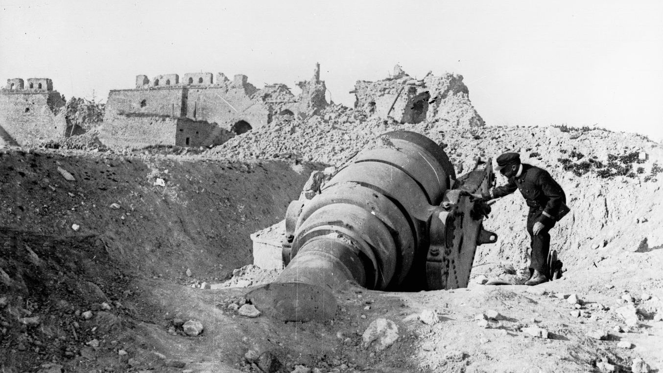 L'image montre un homme travaillant près d'un grand canon ou d'une pièce d'artillerie en position, probablement dans un environnement de guerre. À l'arrière-plan, on peut voir des bâtiments en ruine, ce qui suggère que la scène se déroule dans une zone de conflit, possiblement après un bombardement. Le paysage est aride et rempli de débris, ce qui accentue l'atmosphère de destruction. L'homme semble concentré sur son travail, illustrant les efforts militaires dans un contexte difficile.