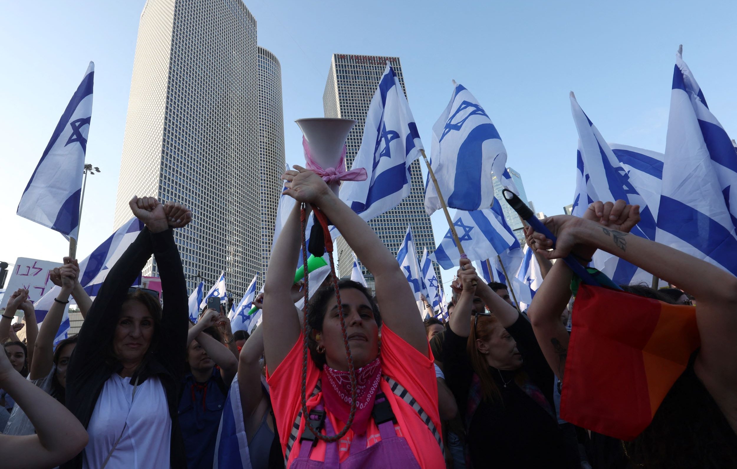 L'image montre une foule de manifestants tenant des drapeaux israéliens. Au centre, une femme portant un haut fluorescent rose élève les bras, tenant un mégaphone. Les manifestants semblent être engagés et passionnés, réunis devant des bâtiments modernes qui dominent l'arrière-plan. L'atmosphère semble être festive et revendicative.