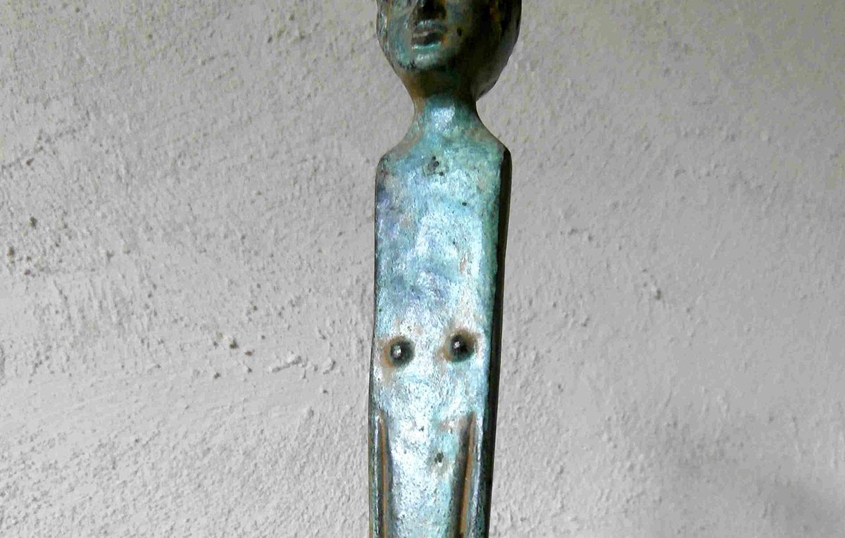 L'image montre un objet en métal, probablement en bronze, qui semble être une sorte de sculpture ou de barre. La partie supérieure représente une tête humaine stylisée, tandis que la tige qui descend présente des éléments qui pourraient évoquer un corps. La surface a une patine verdâtre, indiquant son ancienneté. L'ensemble de l'objet évoque une forme artistique, fusionnant des éléments figuratifs et abstraits.