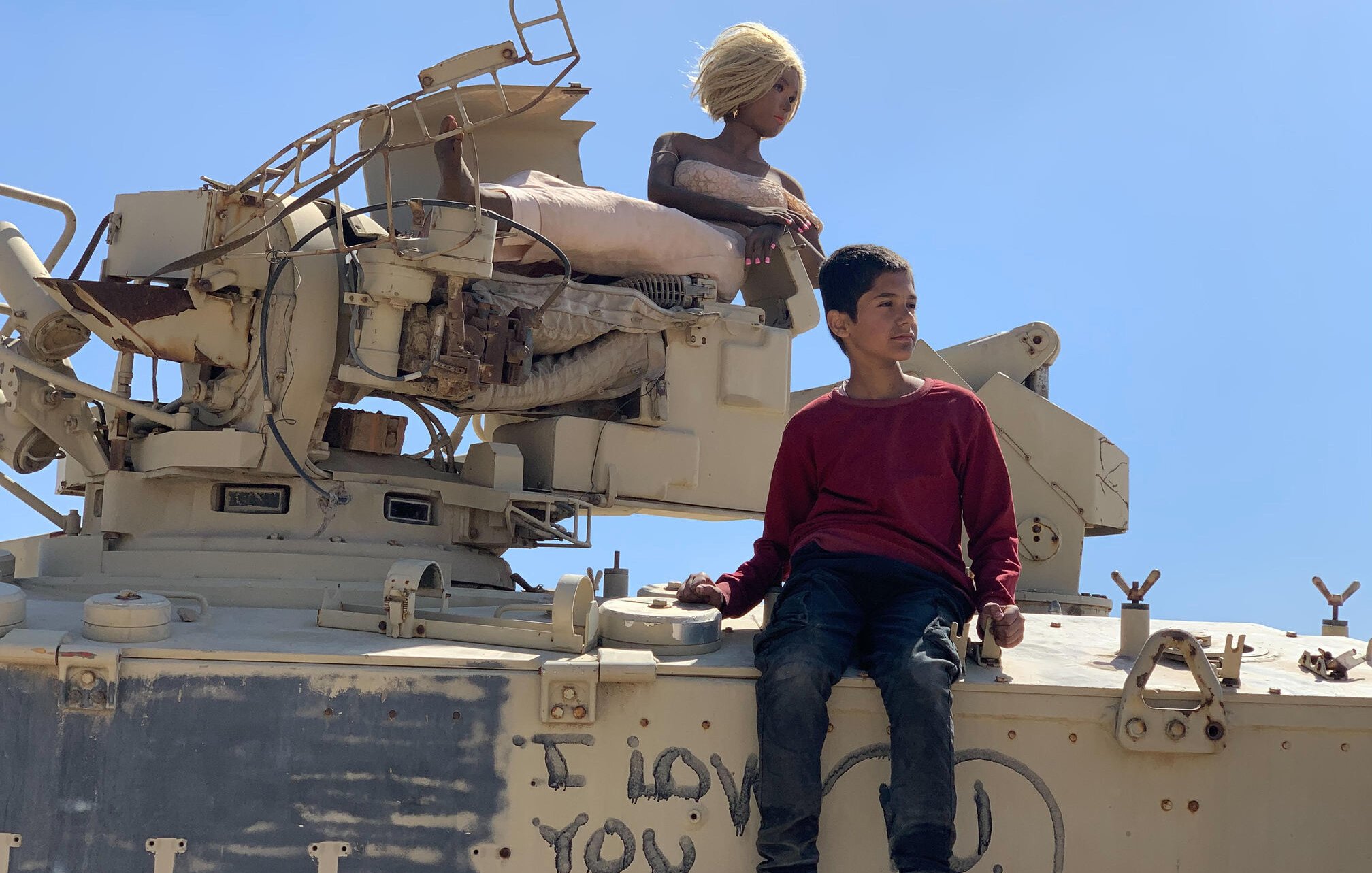 L'image montre un garçon assis sur un véhicule militaire. À côté de lui, il y a une poupée aux cheveux blonds, perchée sur le véhicule. Le fond est clair, avec un ciel bleu. Sur le véhicule, on peut voir des inscriptions, dont "I LOVE YOU" en lettres. L'ensemble de la scène dégage une atmosphere contrastée entre l'innocence de l'enfant et le décor militaire.