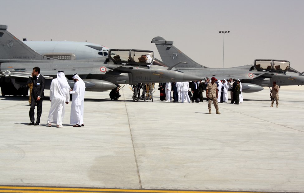 L'image montre une scène sur un tarmac d'aéroport militaire. On y voit plusieurs avions de chasse, probablement des Mirage ou des jets similaires, garés sur une surface bétonnée. Des membres d'équipage, habillés en uniforme, interagissent avec des personnes portant des costumes traditionnels. En arrière-plan, il y a d'autres personnes rassemblées, ce qui suggère un événement ou une cérémonie. Le cadre est désert, avec un ciel clair, ce qui indique qu'il pourrait s'agir d'une région aride.