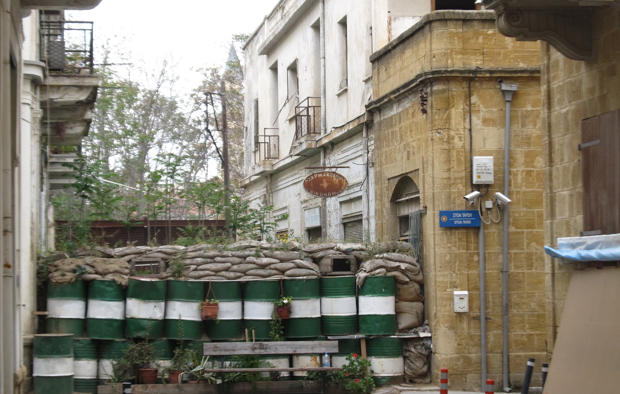 L'image montre une rue étroite avec un mur constitué de sacs de sable et de fûts en plastique, probablement utilisés comme une barrière de protection. Au fond, il y a des bâtiments anciens avec des balcons, et des signes de végétation qui poussent sur le mur. L'atmosphère de la scène semble être à la fois urbaine et quelque peu abandonnée, avec un mélange de vie naturelle et d'éléments de construction.