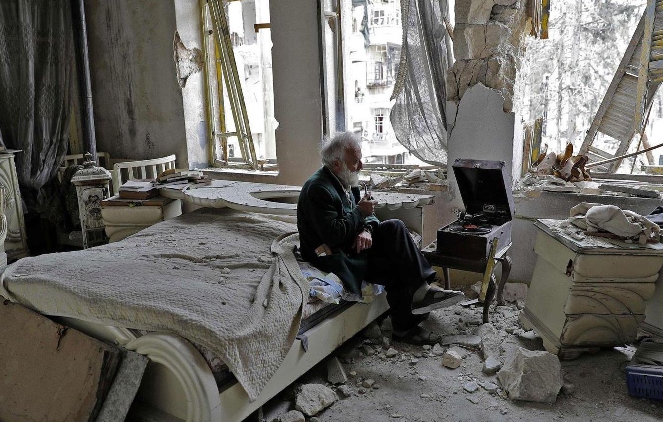 L'image montre un espace intérieur dévasté, probablement un appartement. Au centre, un homme âgé est assis sur un lit, tenant un objet à la main, peut-être une cigarette. L'environnement est marqué par des murs endommagés, des débris éparpillés et des fenêtres brisées, laissant voir l'extérieur. La lumière naturelle pénètre par les ouvertures, révélant l'état de désolation de la pièce. L'ambiance est sombre et mélancolique, illustrant les conséquences d'un conflit ou d'une catastrophe.