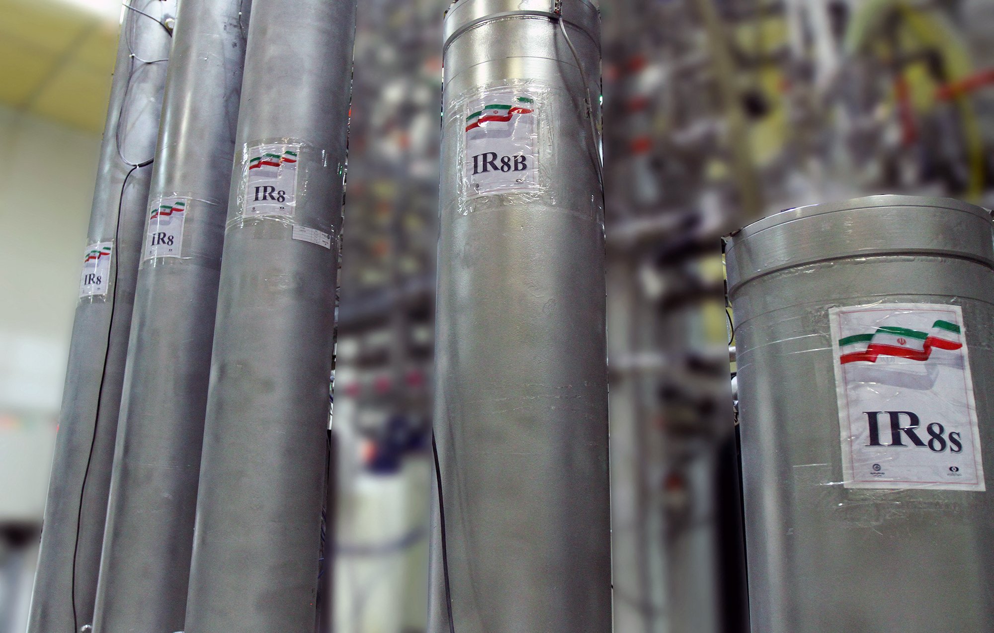 L'image montre plusieurs cylindres métalliques étiquetés avec des inscriptions visibles, y compris "IR8" et "IR8b". Ces cylindres semblent faire partie d'une installation technique, probablement liée à l'enrichissement de l'uranium, étant donné leur apparence et le contexte. En arrière-plan, on peut apercevoir d'autres équipements complexes, suggérant un environnement industriel ou scientifique avancé. Les étiquettes affichent également des drapeaux stylisés, probablement en référence à un pays spécifique.