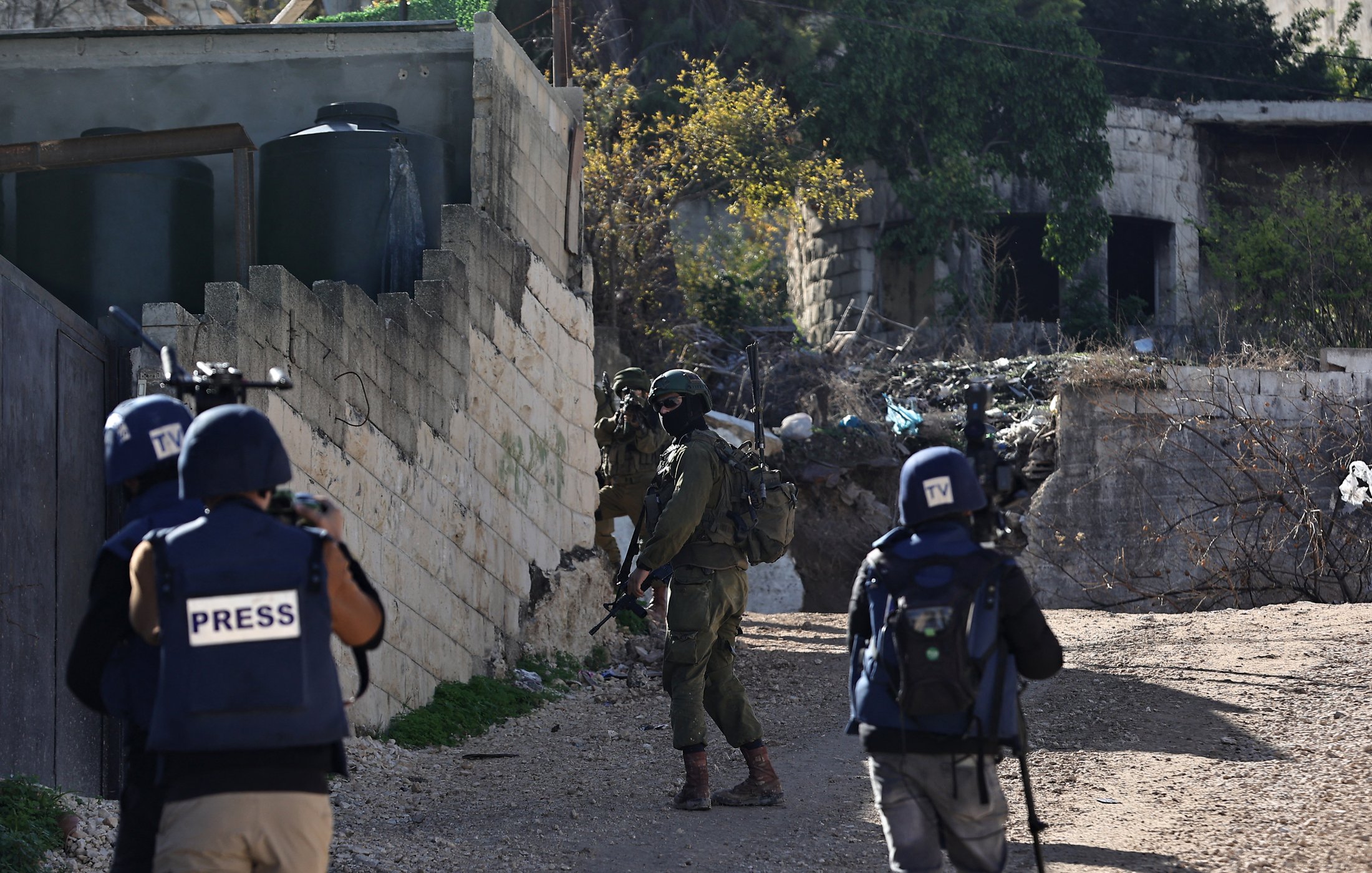 L'image montre une scène de conflit avec des soldats armés et des journalistes. Il y a des soldats en uniforme, portant des casques et des équipements de protection, qui se déplacent dans un environnement urbain, probablement dans une zone de conflit. Des journalistes sont également présents, identifiables grâce à leurs gilets marqués du mot "PRESS". En arrière-plan, on peut apercevoir des structures en ruine et des éléments de débris, ce qui suggère un contexte de tensions ou de violence.