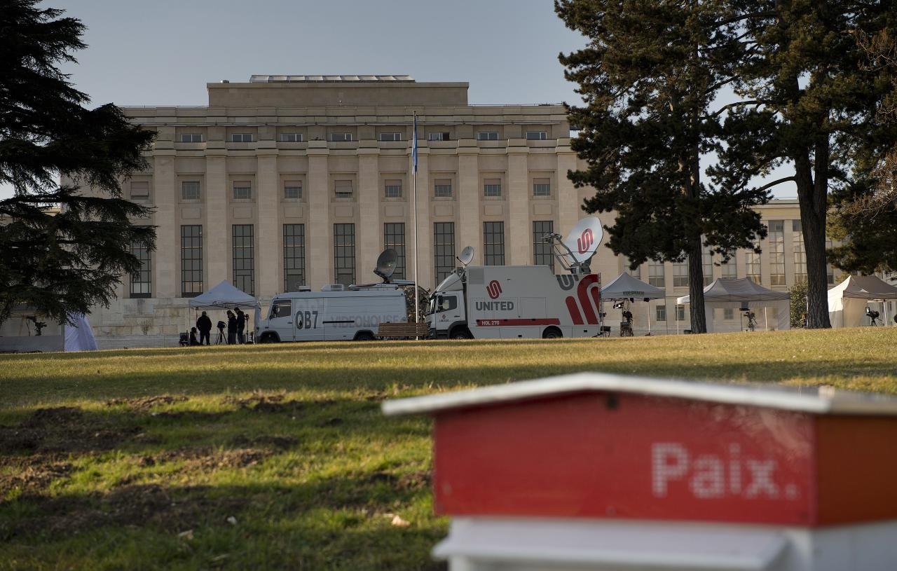 L'image montre un bâtiment imposant, probablement un palais ou un bâtiment gouvernemental, situé à Genève. À l'avant-plan, on aperçoit une petite structure rouge portant le mot "Paix". En arrière-plan, on distingue des véhicules de télévision et des tentes, indiquant qu'un événement ou une conférence pourrait avoir lieu. Le cadre est agrémenté d'arbres, suggérant un espace extérieur agréable.