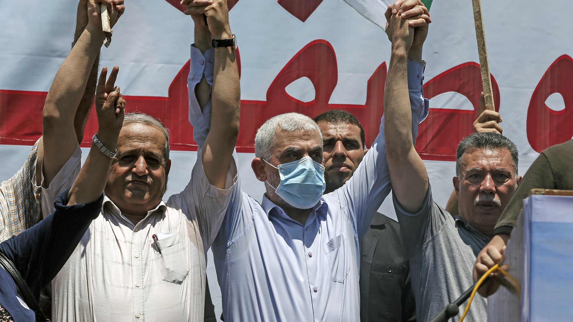 L'image montre un groupe de personnes levant les bras en signe de solidarité ou de protestation. Certains portent des masques, et il y a des drapeaux visibles en arrière-plan. L'ambiance semble être celle d'un rassemblement ou d'une manifestation. Les expressions des participants varient, reflétant des émotions de détermination ou de franche camaraderie.