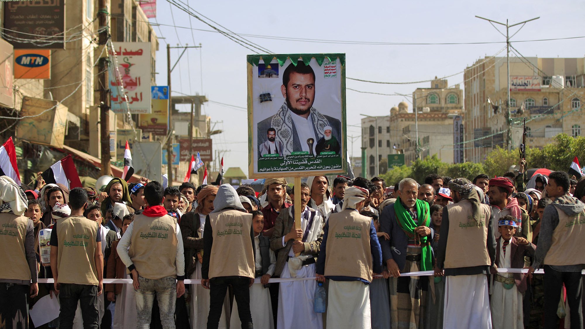 L'image montre une manifestation dans une rue animée, avec une foule de personnes rassemblées. Au premier plan, il y a un groupe de manifestants portant des vêtements traditionnels et des gilets beige. Ils se tiennent derrière une barrière et semblent faire face à l'objectif. En arrière-plan, on voit des drapeaux, ainsi qu'une grande affiche représentant une personne, probablement un leader ou une figure importante. L'ambiance semble être chargée d'émotion et de détermination, avec un secteur urbain typique visible autour.