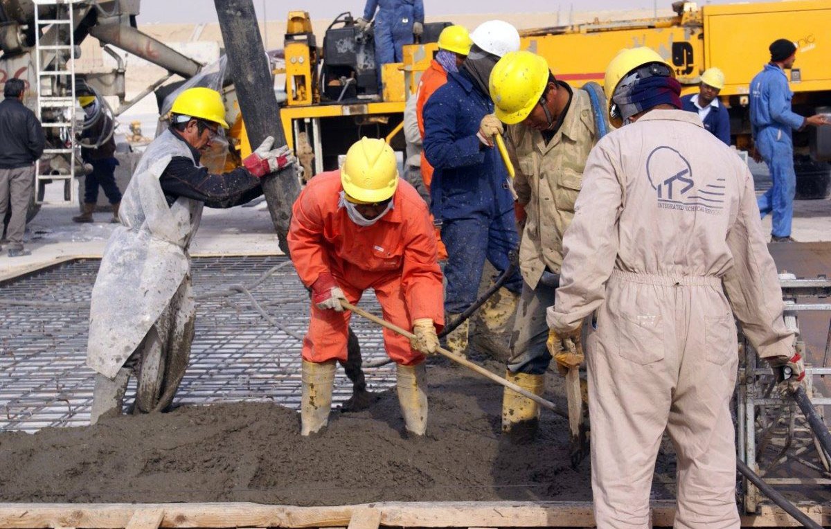 L'image montre plusieurs ouvriers en train de travailler sur un chantier de construction. Ils portent des casques de sécurité jaunes et des vêtements de protection, tels que des combinaisons et des bottes en caoutchouc. L'un des travailleurs utilise une pelle pour manipuler du béton, tandis que d'autres semblent s'occuper de l'installation ou de la préparation du béton. En arrière-plan, on peut voir des machines de chantier et des équipements. L'atmosphère est active, indiquant un environnement de travail intense.