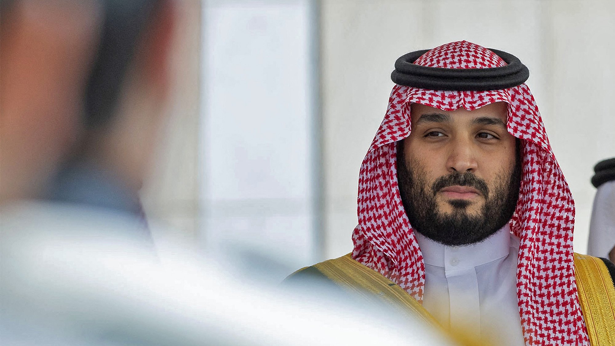 L'image montre un homme portant des vêtements traditionnels saoudiens, y compris un keffieh et un thobe. Il a une expression sérieuse et regarde vers la caméra ou un point de vue adjacent. Son entourage semble formel, avec une ambiance de cérémonie ou de rencontre officielle. Les couleurs dominantes sont les tons de rouge et de blanc du keffieh, ainsi que des touches dorées sur sa tenue.