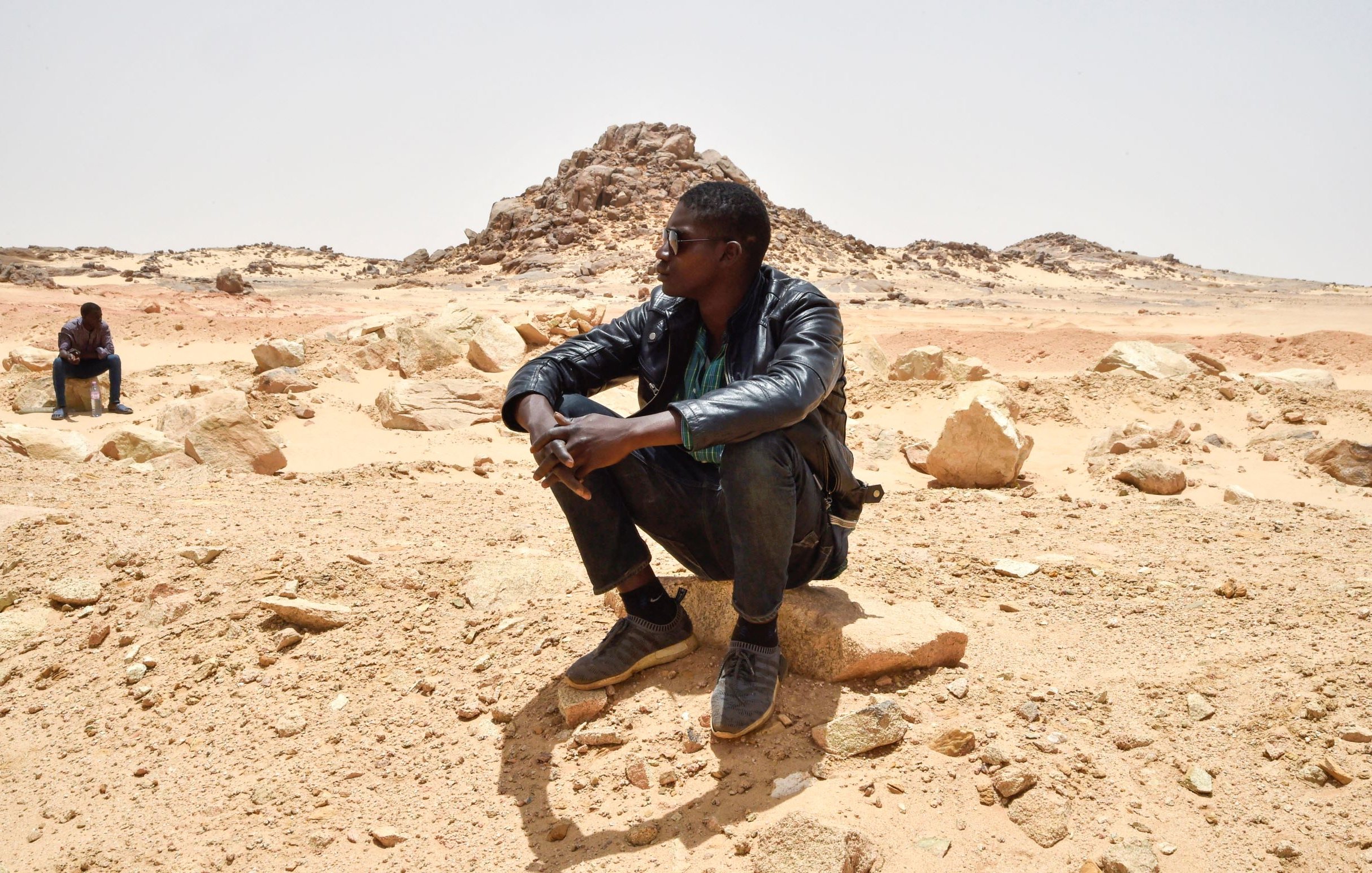 L'image montre deux personnes assises dans un paysage désertique. L'un des hommes est en train de s'asseoir sur une grosse pierre, portant une veste en cuir et des lunettes de soleil. Il semble pensif, observant le paysage autour de lui. En arrière-plan, une autre personne est également assise, mais davantage éloignée. Le terrain est constitué de sable et de roches, avec des formations rocheuses au loin, sous un ciel clair. L'atmosphère semble calme et isolée.