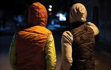 L'image montre deux personnes de dos, se tenant à proximité l'une de l'autre. L'une porte un gilet d'une couleur orange avec des manches vertes, tandis que l'autre est vêtu d'un gilet noir et d'une capuche grise. Ils semblent se trouver dans une rue sombre, avec quelques lumières au loin, ce qui donne une ambiance nocturne à la scène. L'une des personnes pointe quelque chose du doigt, suggérant une conversation ou une observation partagée.