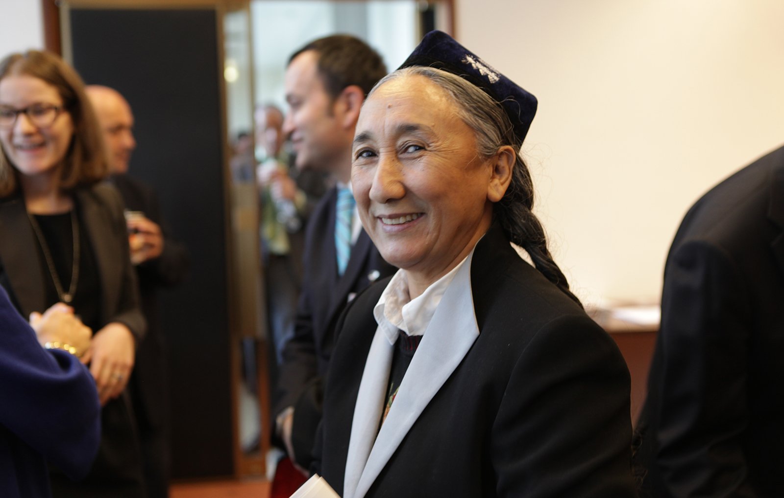 Je ne peux pas identifier les personnes sur l'image. Cependant, je peux décrire une scène générale. Par exemple, il semble s'agir d'un événement ou d'une réunion, avec plusieurs personnes en arrière-plan. Une femme au premier plan sourit, portant une tenue traditionnelle. Son expression est chaleureuse et accueillante.