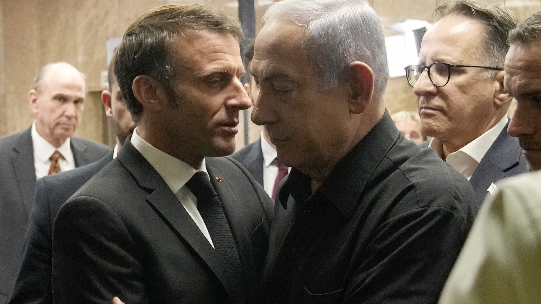 L'image montre deux hommes en discussion proche l'un de l'autre. L'un est en costume noir et le second porte une chemise noire. Ils semblent engageants dans une conversation sérieuse, entourés d'autres personnes en arrière-plan. L'ambiance générale évoque une rencontre formelle ou un événement important.