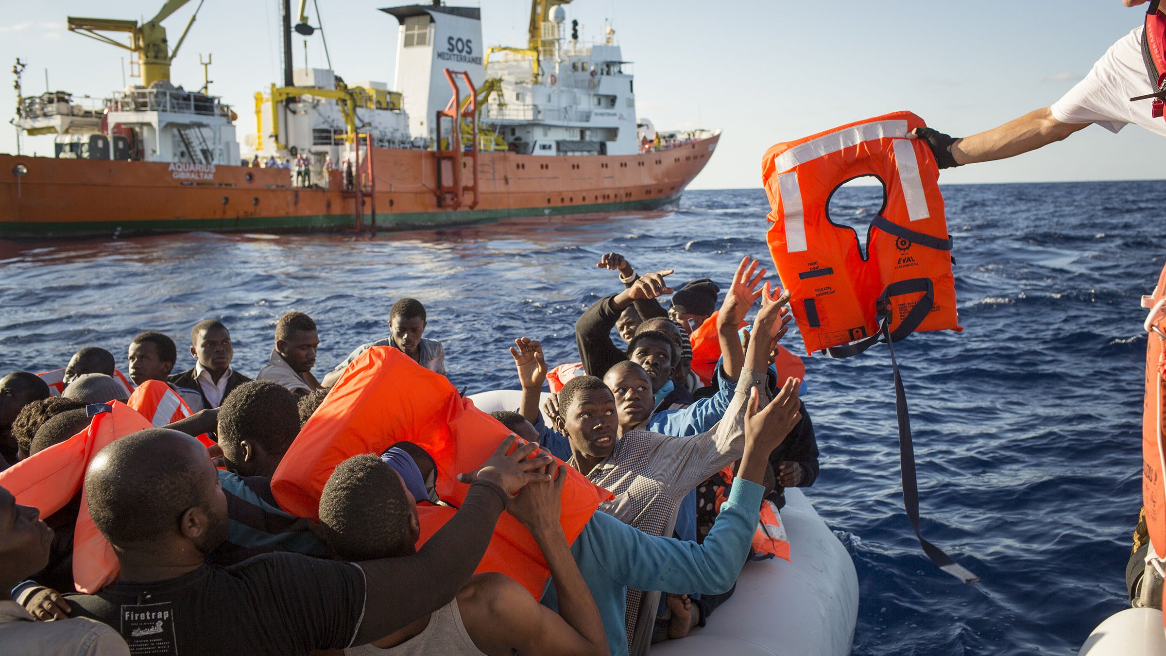 L'image montre une scène en mer où un groupe de personnes se trouve sur un canot pneumatique. Elles semblent en détresse et soulèvent les bras en direction d'un sauveteur qui leur tend une bouée de sauvetage. En arrière-plan, un grand navire de secours est visible, avec des couleurs orange et blanc. L'atmosphère semble tendue, et on peut ressentir l'urgence de la situation. Le ciel est dégagé et l'eau est calme, contrastant avec la gravité du moment.