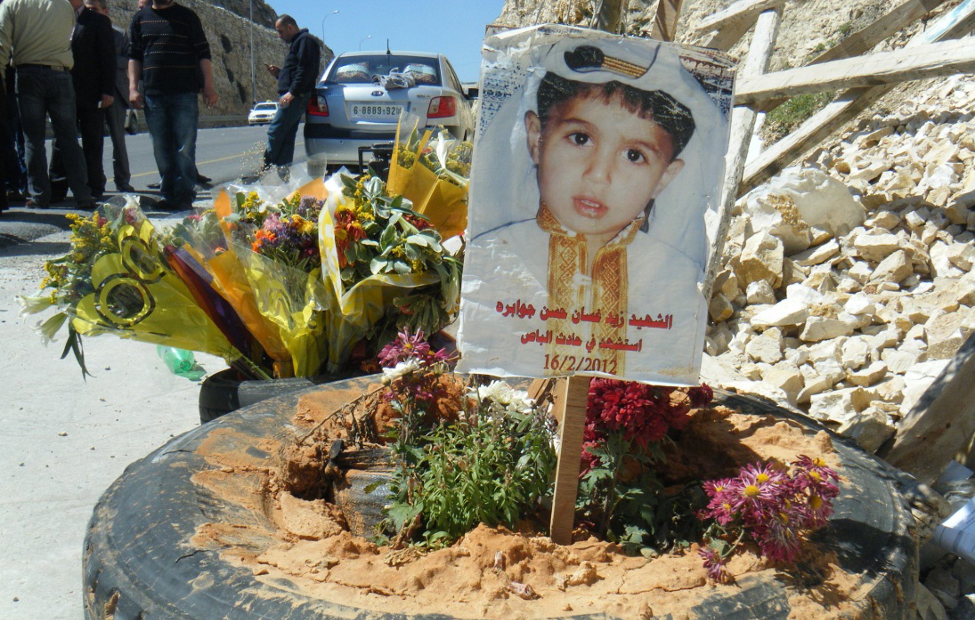 L'image montre un mémorial pour un enfant, avec une photo de lui affichée sur un panneau en bois planté dans un pneu. Autour du panneau, il y a des fleurs multicolores et de la terre. En arrière-plan, on peut voir des personnes rassemblées et un paysage de pierre. Cela semble être un hommage émouvant à un enfant décédé.