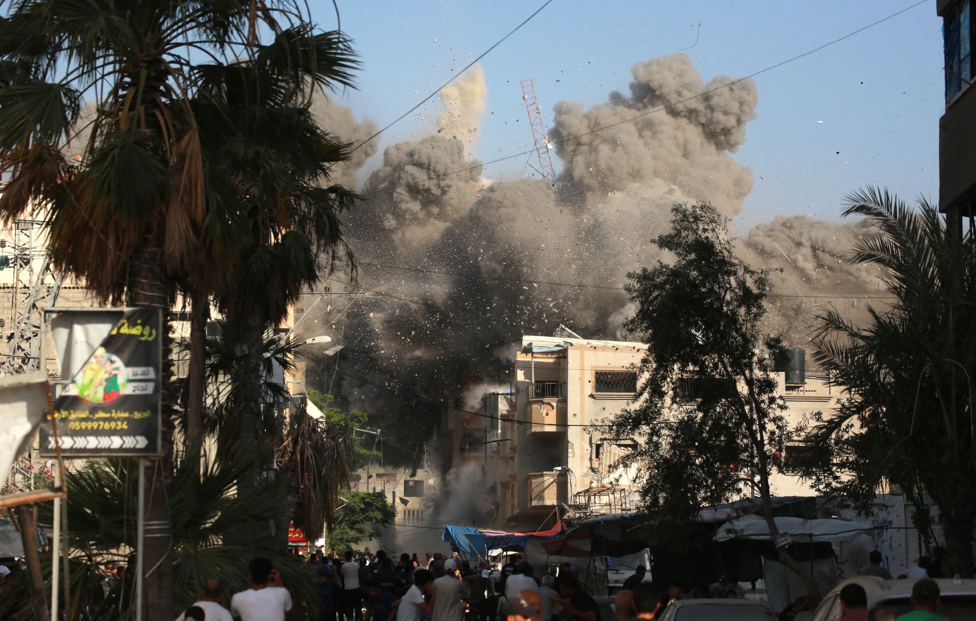 L'image montre une scène chaotique en milieu urbain, où des explosions entraînent un nuage de poussière et de débris. Des bâtiments semblent être endommagés, créant un environnement de destruction. On peut voir des gens dans la rue, réagissant à la situation, ainsi que des palmiers qui ajoutent une touche de verdure au paysage. Le ciel est clair, en contraste avec la fumée épaisse et sombre qui s'élève des ruines.