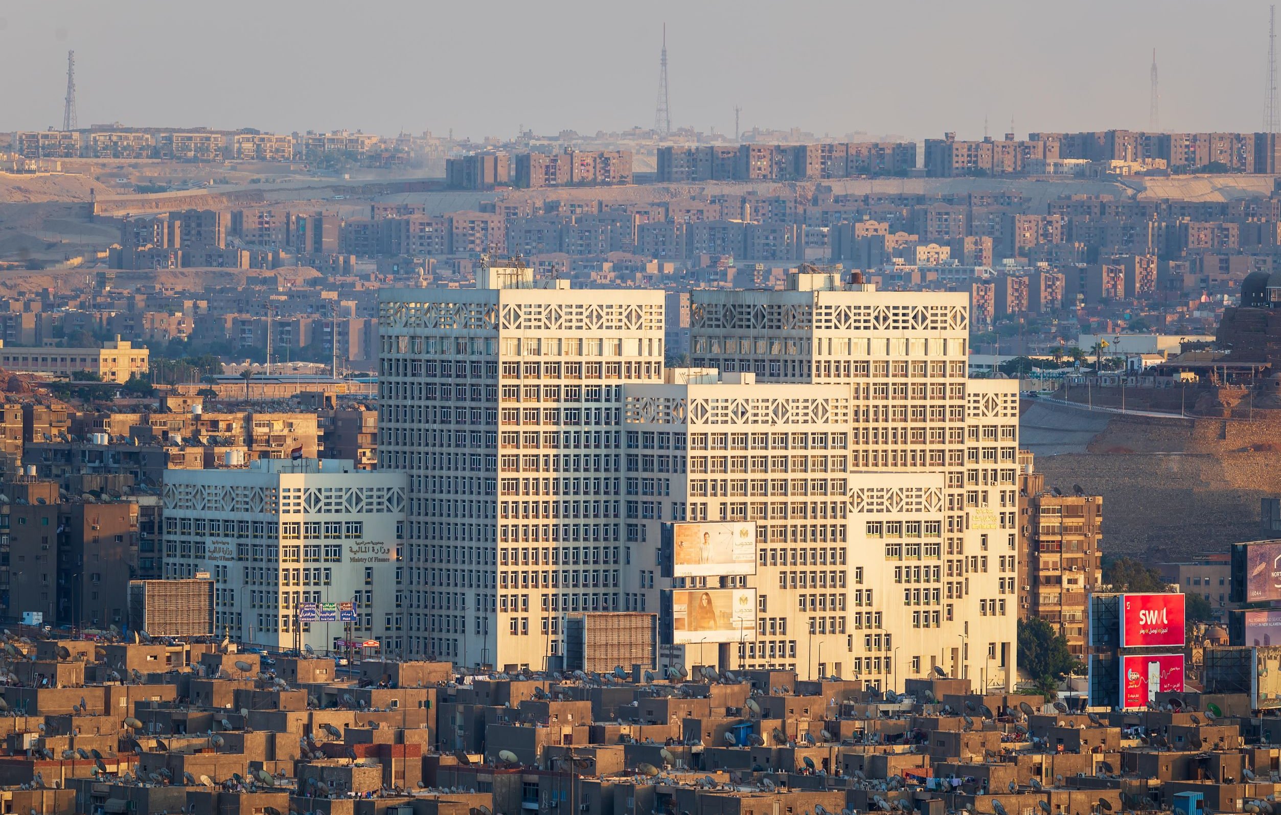 L'image montre un paysage urbain avec plusieurs bâtiments modernes au premier plan, qui se distinguent par leur architecture unique et leurs façades vitrées. En arrière-plan, on aperçoit une vaste étendue de constructions résidentielles plus anciennes, avec des habitations compactes et souvent uniformes. La lumière du soleil semble éclairer les bâtiments modernes, créant un contraste avec le reste de la ville, particulièrement à l'heure dorée du matin ou du soir. Le ciel apparaît clair, ce qui met en valeur la scène urbaine.