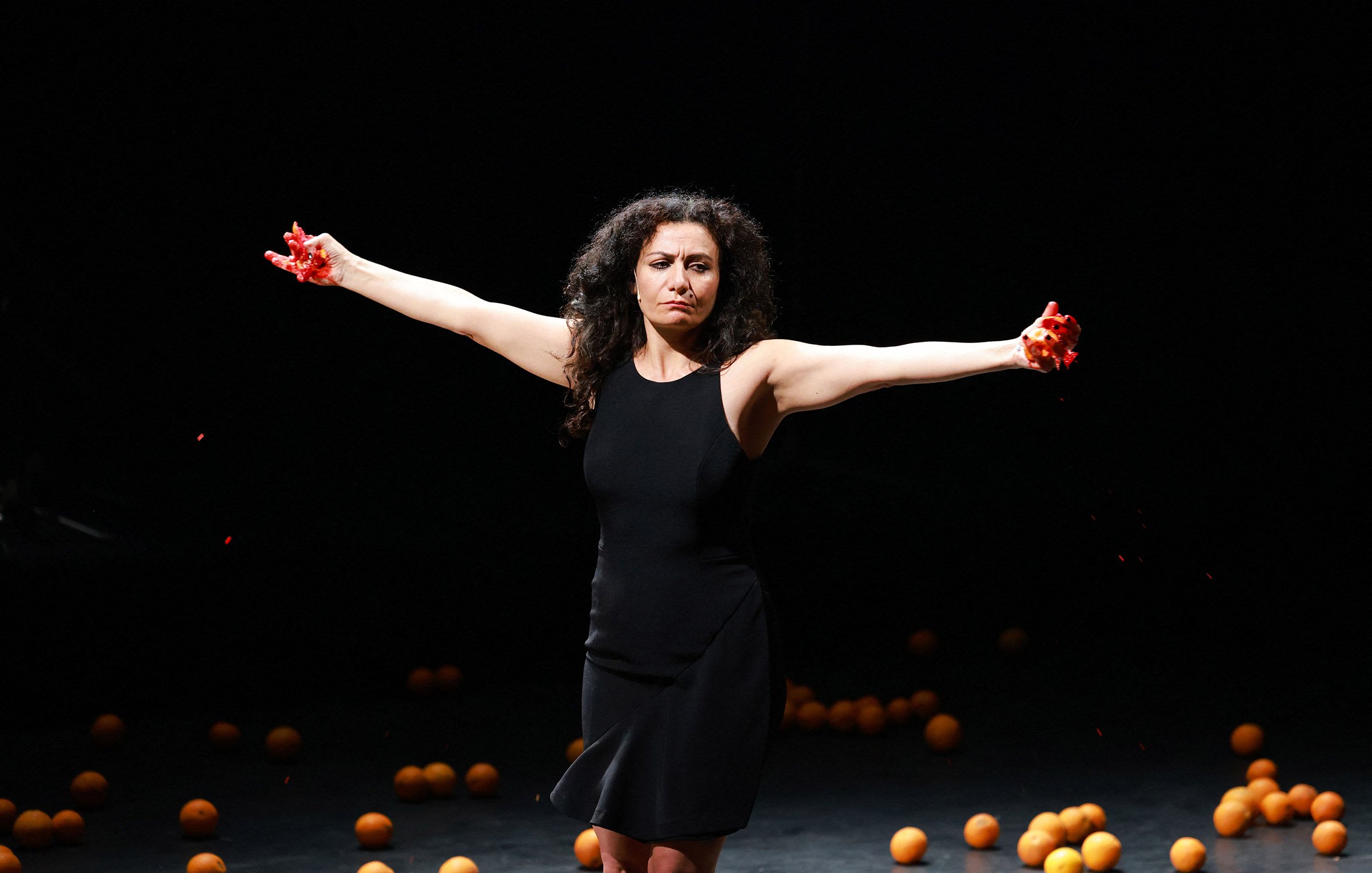 L'image montre une femme debout sur scène, les bras étendus de chaque côté. Elle porte une robe noire et exprime une émotion intense. Des oranges sont dispersées autour d'elle sur le sol, ajoutant une dimension visuelle frappante à la scène. Des éclaboussures de liquide rouge semblent provenir de ses mains, ce qui accentue le drame et la tension de l'instant. L'éclairage de la scène est sombre, mettant ainsi en valeur la silhouette de la femme et les oranges.