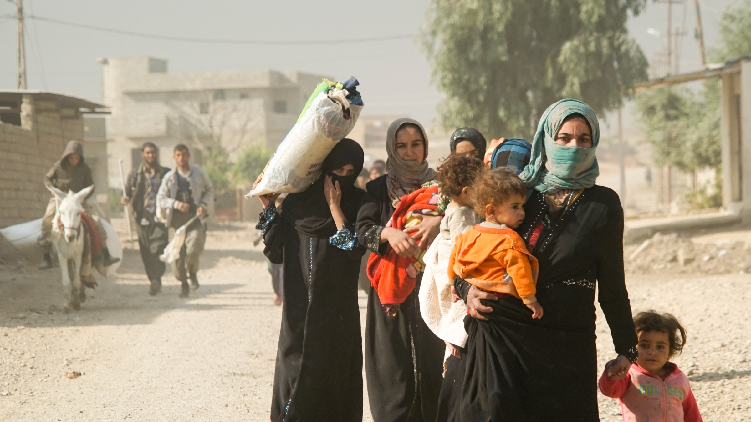 L'image montre un groupe de femmes et d'enfants marchant sur un chemin en terre. Certaines femmes portent des vêtements traditionnels et des foulards. L'une d'elles porte un enfant sur son bras, tandis qu'une autre porte un paquet sur sa tête. En arrière-plan, on peut voir des bâtiments et une personne à cheval s'approchant. L'atmosphère semble poussiéreuse et le paysage est rural. Les expressions des femmes sont variées, allant de la concentration à la sollicitude envers les enfants.