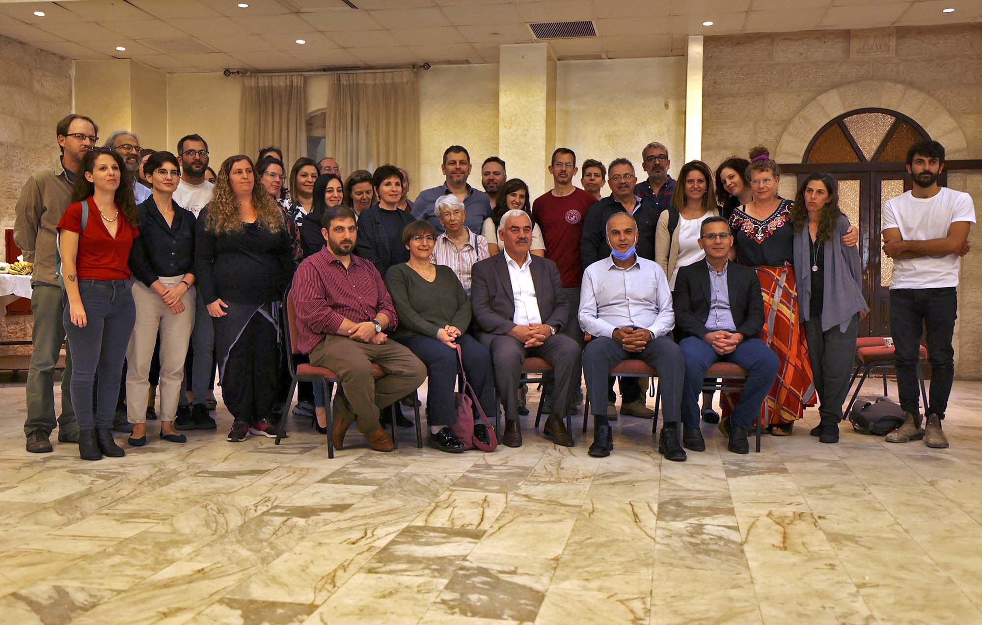L'image montre un grand groupe de personnes rassemblées dans une salle, probablement pour un événement ou une célébration. Les gens sont assis et debout, souriant et regardant vers l'objectif. L'espace présente des murs en pierre et un sol en marbre, créant une atmosphère conviviale. On peut remarquer une variété de styles vestimentaires parmi les participants, ce qui indique une diversité au sein du groupe.