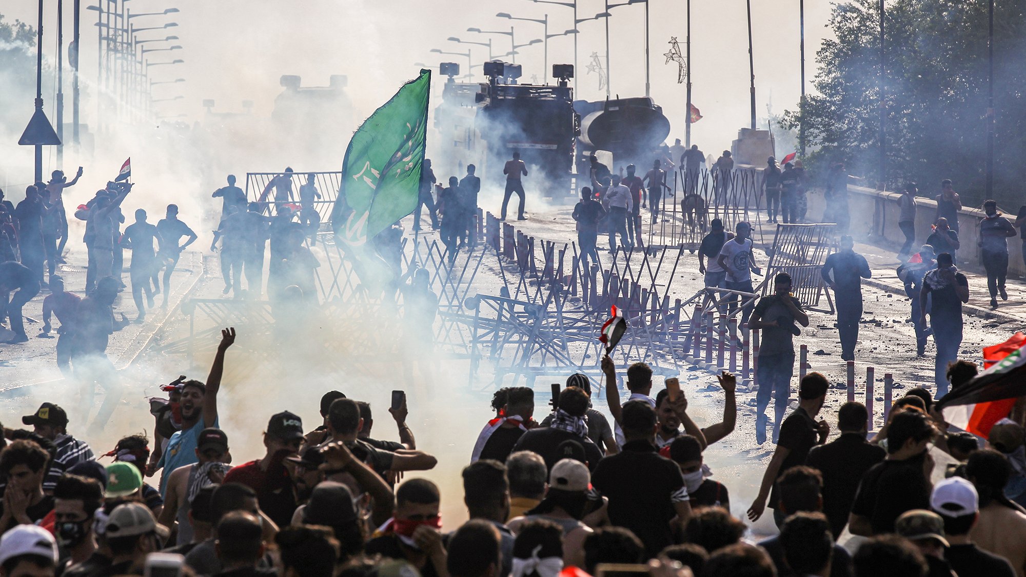 L'image montre une scène de tumultes ou de manifestations. On y voit un grand groupe de personnes rassemblées, brandissant des drapeaux et levant les bras. Il y a des barrages en bois et de la fumée, probablement causée par des gaz lacrymogènes. À l'arrière-plan, des véhicules blindés sont visibles, renforçant l'idée d'une situation tendue. L'atmosphère est chargée et chaotique, avec des manifestants qui semblent déterminés et en mouvement.