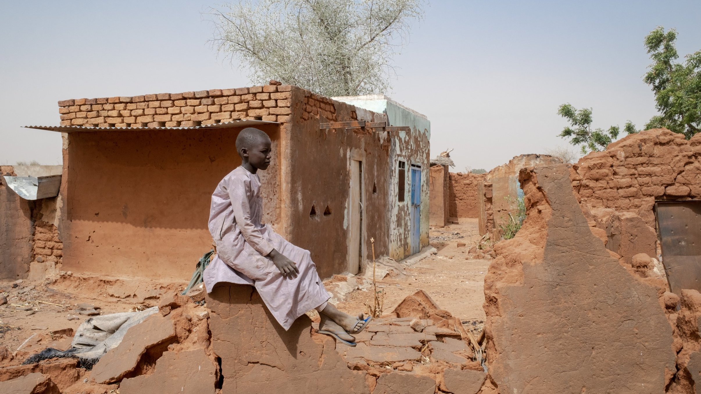 L'image montre un jeune garçon assis sur de décombres dans une région désertique. Il porte une tunique traditionnelle et semble pensif. En arrière-plan, il y a des bâtiments en terre cuite, dont certains sont partiellement détruits. Le paysage est aride, avec quelques arbres éparpillés, et le ciel est clair. L'atmosphère dégage une impression de solitude et de résilience face aux conditions difficiles.