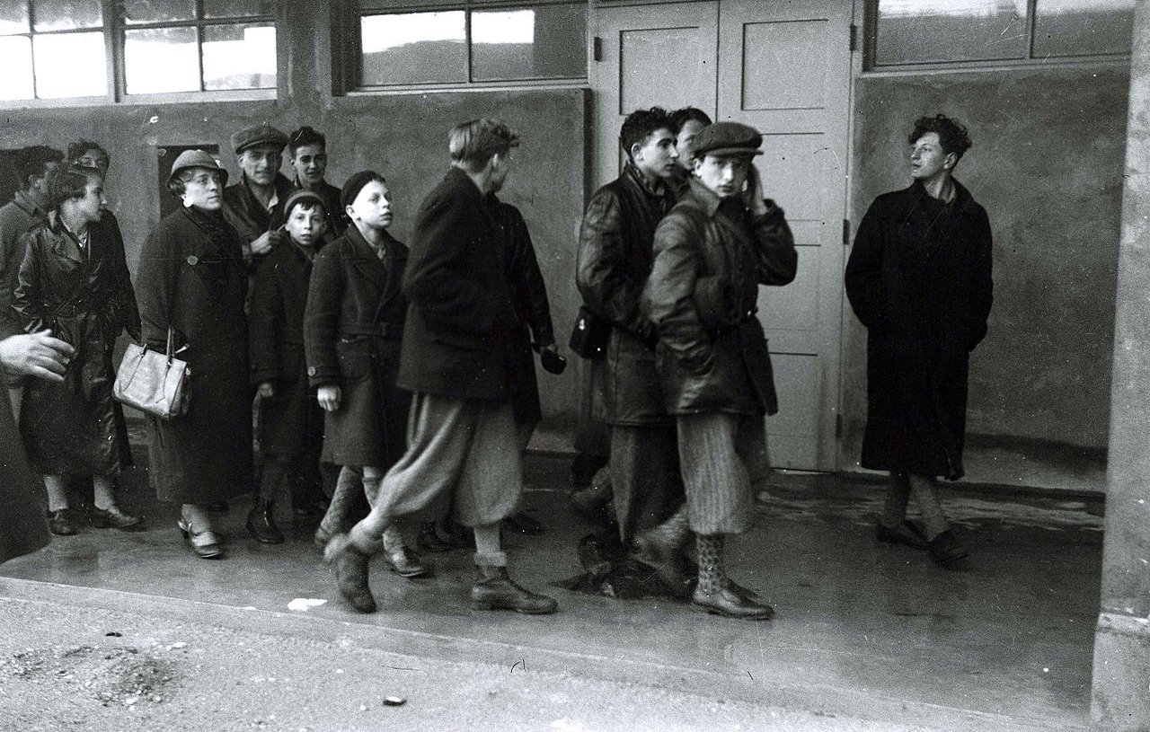 L'image montre un groupe de personnes, majoritairement des femmes et des enfants, marchant en direction d'un bâtiment. Ils portent des vêtements sombres et d'hiver. Leurs expressions semblent sérieuses et préoccupées. À l'entrée du bâtiment, une personne se tient à l'écart, observant le groupe avec une expression indéchiffrable. L'atmosphère de l'image est pesante et évoque un sentiment de tristesse ou d'incertitude. La scène semble avoir lieu dans un cadre urbain ou un lieu de rassemblement, avec des fenêtres en arrière-plan.