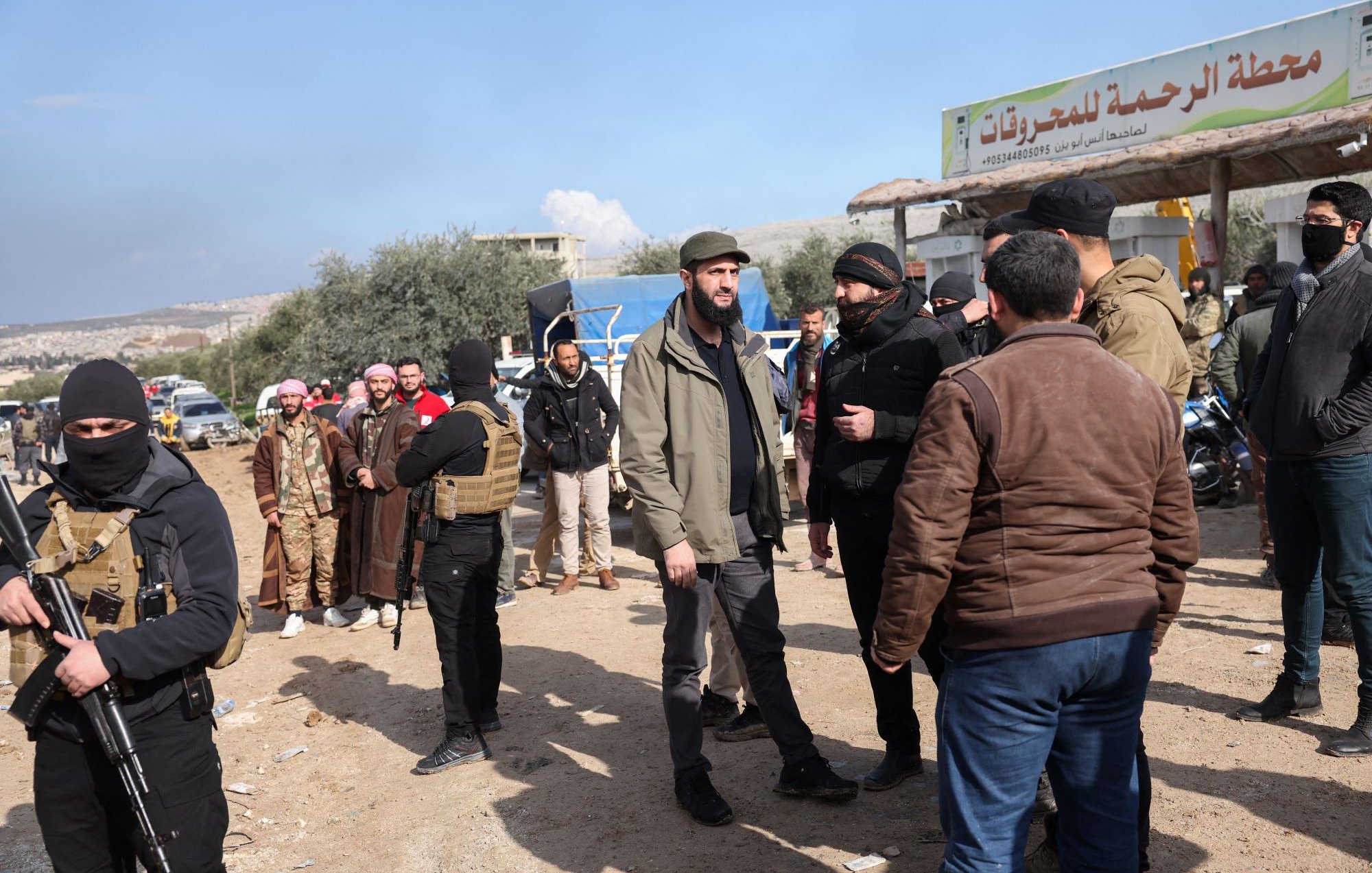 L'image montre un groupe de personnes réunies dans un environnement en plein air, vraisemblablement dans une zone rurale ou semi-urbaine. Certains hommes portent des vêtements traditionnels, tandis que d'autres sont habillés de manière plus moderne. Il y a également des individus en tenue de camouflage avec des gilets tactiques, qui semblent être armés. En arrière-plan, on peut apercevoir un panneau signalétique. Le ciel est partiellement nuageux, ce qui suggère une atmosphère de journée. Les gens interagissent entre eux, ce qui laisse penser qu'il s'agit d'une rencontre ou d'une discussion.