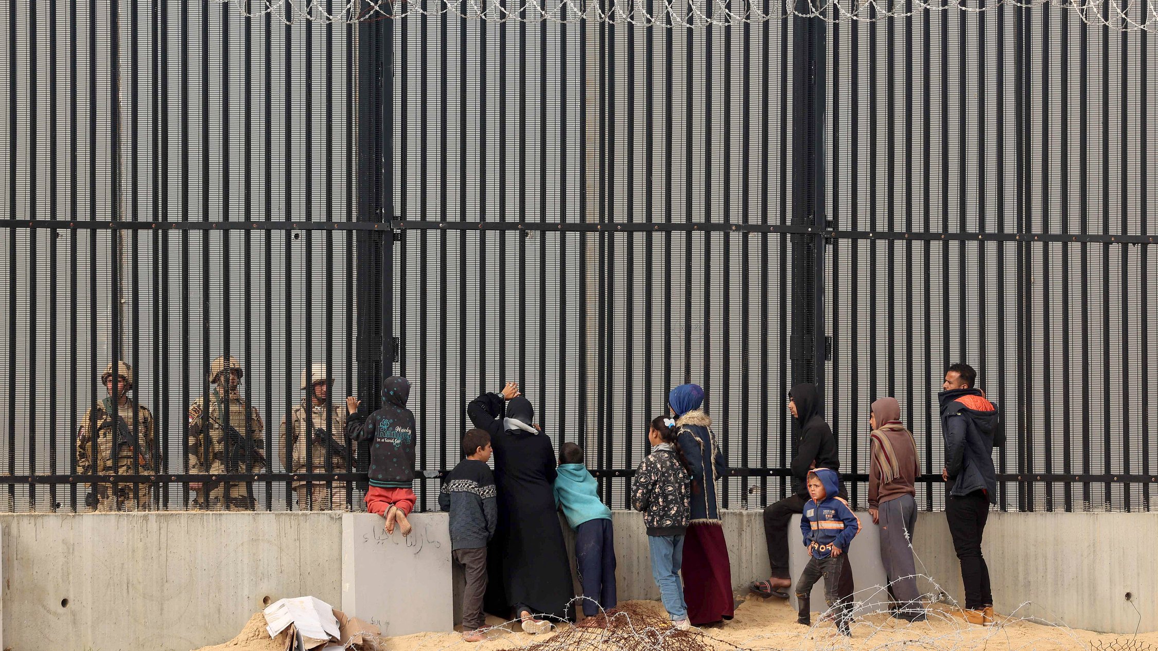 L'image montre un groupe de personnes se tenant devant une grande clôture métallique, probablement une barrière de sécurité. De l'autre côté, des soldats en uniforme semblent observer le groupe. Les personnes sont composées de familles et d'enfants, certains d'eux essaient de communiquer ou de se rapprocher de la clôture. Le cadre est marqué par un environnement désertique, avec des barbelés visibles au sol. L'atmosphère semble chargée d'émotion, soulignant une séparation entre les civils et les forces de sécurité.