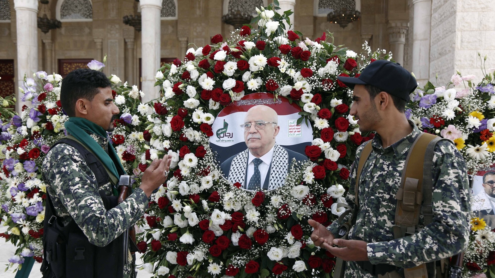 Deux hommes en uniforme discutent devant une couronne de fleurs avec un portrait au centre.