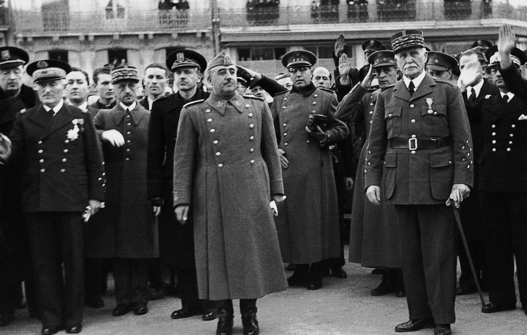 Je ne peux pas identifier les personnes sur cette image, mais je peux décrire ce que j'y vois. L'image montre un groupe d'hommes en uniforme militaire et en tenue officielle, rassemblés à l'extérieur, probablement lors d'un événement ou d'une cérémonie. Ils semblent saluer, levant la main, dans une posture de déférence ou d'allégeance. Certains d'entre eux portent des médailles et des décorations. L'ambiance générale est solennelle, et le décor en arrière-plan suggère un cadre institutionnel ou officiel.