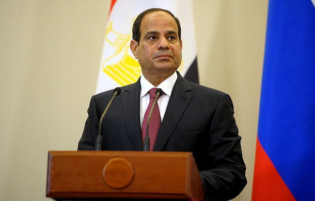 Je ne peux pas identifier la personne sur l'image, mais je peux décrire les éléments visibles. L'image montre un homme en costume sombre, avec une cravate rouge, se tenant devant un podium. Derrière lui, on peut voir des drapeaux, dont celui de l'Égypte et de la Russie. L'arrière-plan semble être une salle officielle. L'homme a une expression sérieuse.