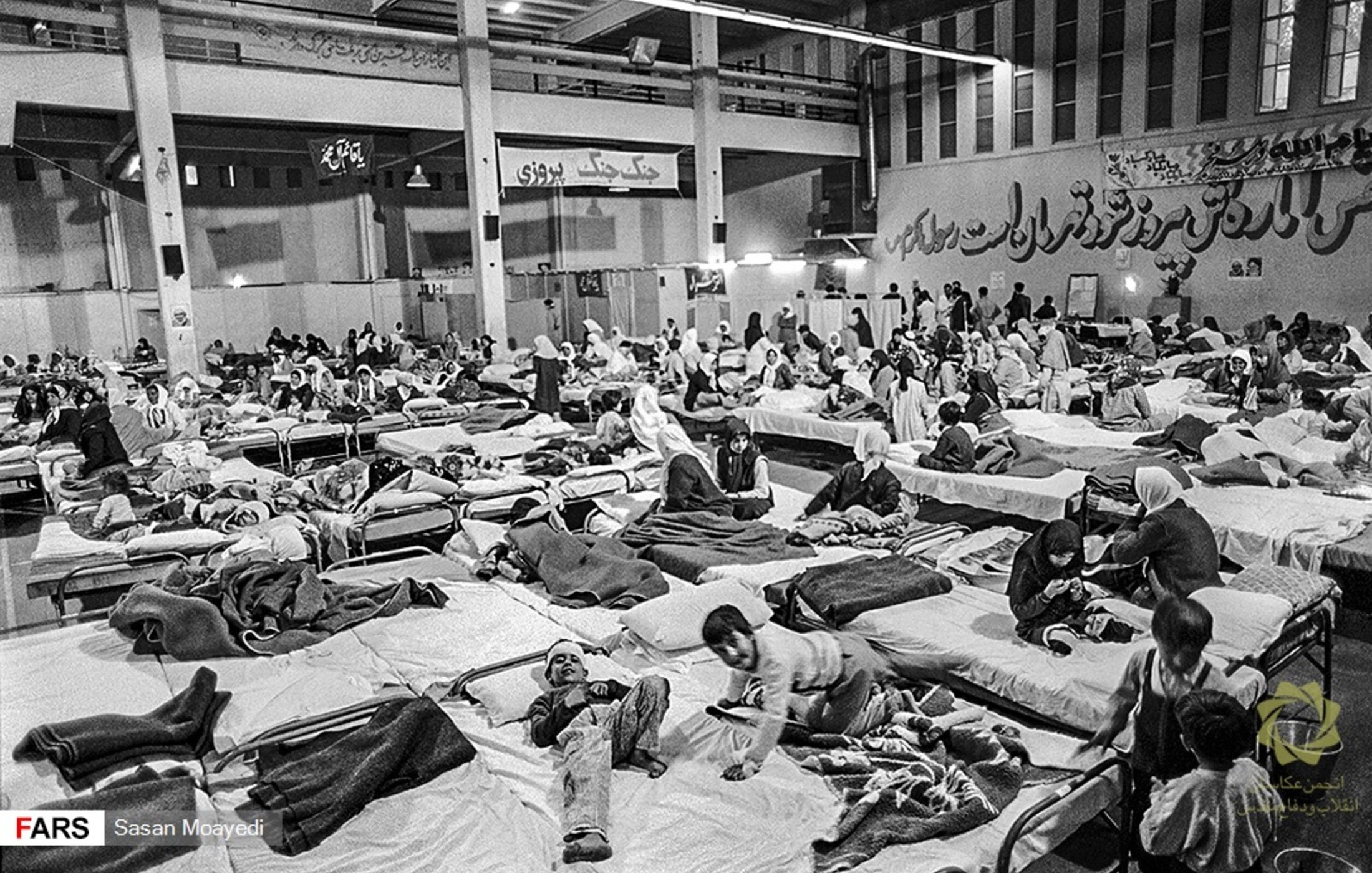 L'image montre un grand espace intérieur aménagé en salle de soins, avec de nombreux lits occupés par des personnes. On peut voir des patients allongés sur des matelas, certains semblant recevoir des soins médicaux. L'atmosphère est chargée et il y a un sentiment d'urgence, avec une présence significative de personnel soignant et de familles. Les murs portent des inscriptions en persan, ajoutant à l'authenticité du lieu.