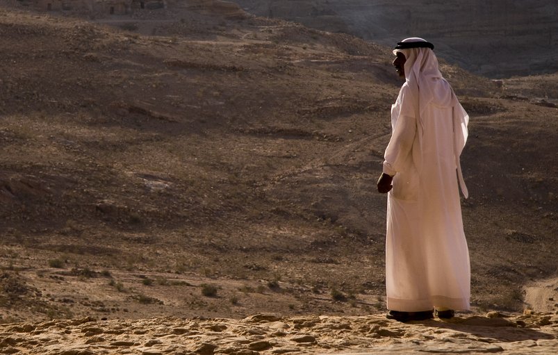 L'image montre une personne vêtue d'une tenue traditionnelle, probablement un kandora ou un dishdasha, se tenant debout sur un terrain désertique. En arrière-plan, on aperçoit un paysage aride avec des collines et des formations rocheuses. La lumière est douce, ce qui donne une ambiance calme et contemplative à la scène. La personne semble regarder au loin, peut-être en train de réfléchir ou d'admirer la vue panoramique du désert.