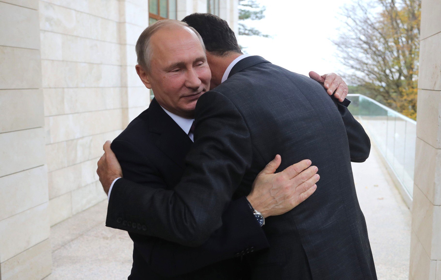 L'image montre deux hommes s'enlaçant dans un cadre officiel. L'un d'eux porte un costume foncé et semble sourire légèrement, tandis que l'autre, également en costume, se penche pour l'étreindre. L'environnement semble être d'une architecture moderne, avec des murs en pierre et un éclairage naturel. L'atmosphère paraît chaleureuse malgré le contexte formel.