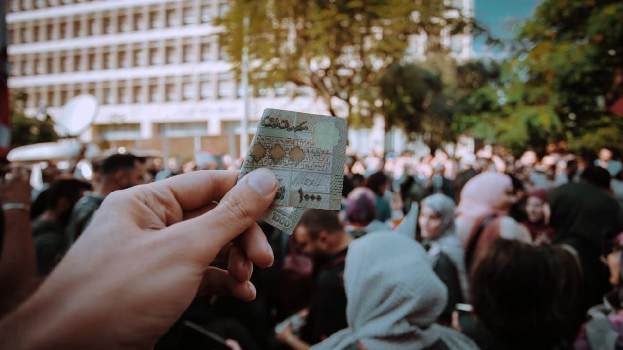 L'image montre une main tenant un billet de banque marocain au premier plan, tandis qu'à l'arrière-plan, une foule est rassemblée. On aperçoit des personnes aux tenues variées, certaines portant des foulards. Le cadre semble être un événement public, probablement une manifestation ou une célébration, avec des bâtiments modernes en arrière-plan et des arbres qui ajoutent une touche de verdure à la scène. L'atmosphère semble dynamique et chargée d'énergie collective.