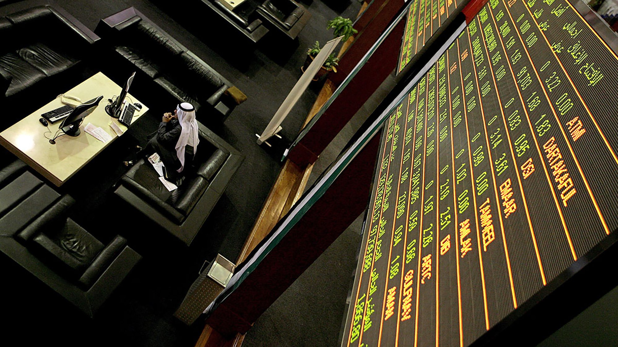 L'image montre un environnement de bourse avec des tables et des sièges confortables. À l'arrière-plan, un grand tableau électronique affiche des informations financières, comme des chiffres et des noms de sociétés. On peut voir quelques personnes, dont un homme en tenue traditionnelle, probablement en train de suivre les fluctuations du marché. L'ambiance semble professionnelle et concentrée, caractéristique d'une salle de négociation.