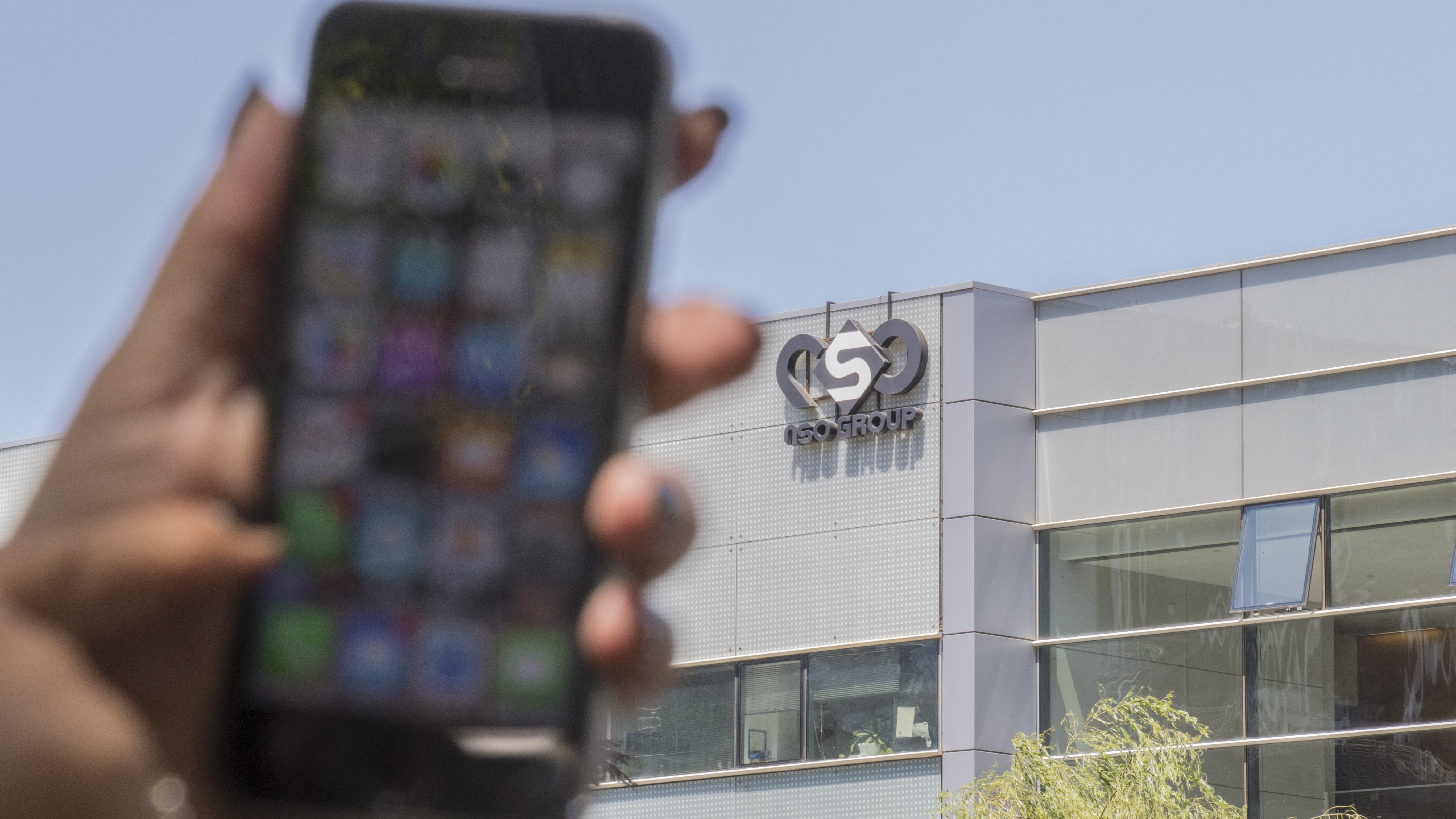 L'image montre un smartphone tenu par une main, flou au premier plan, avec un écran affichant diverses applications. En arrière-plan, on peut voir le bâtiment d'une entreprise, identifié par un logo qui semble être celui d'un groupe nommé "ISN GROUP". Le bâtiment a une façade moderne, probablement en verre. La lumière naturelle éclaire la scène, indiquant qu'il fait jour.