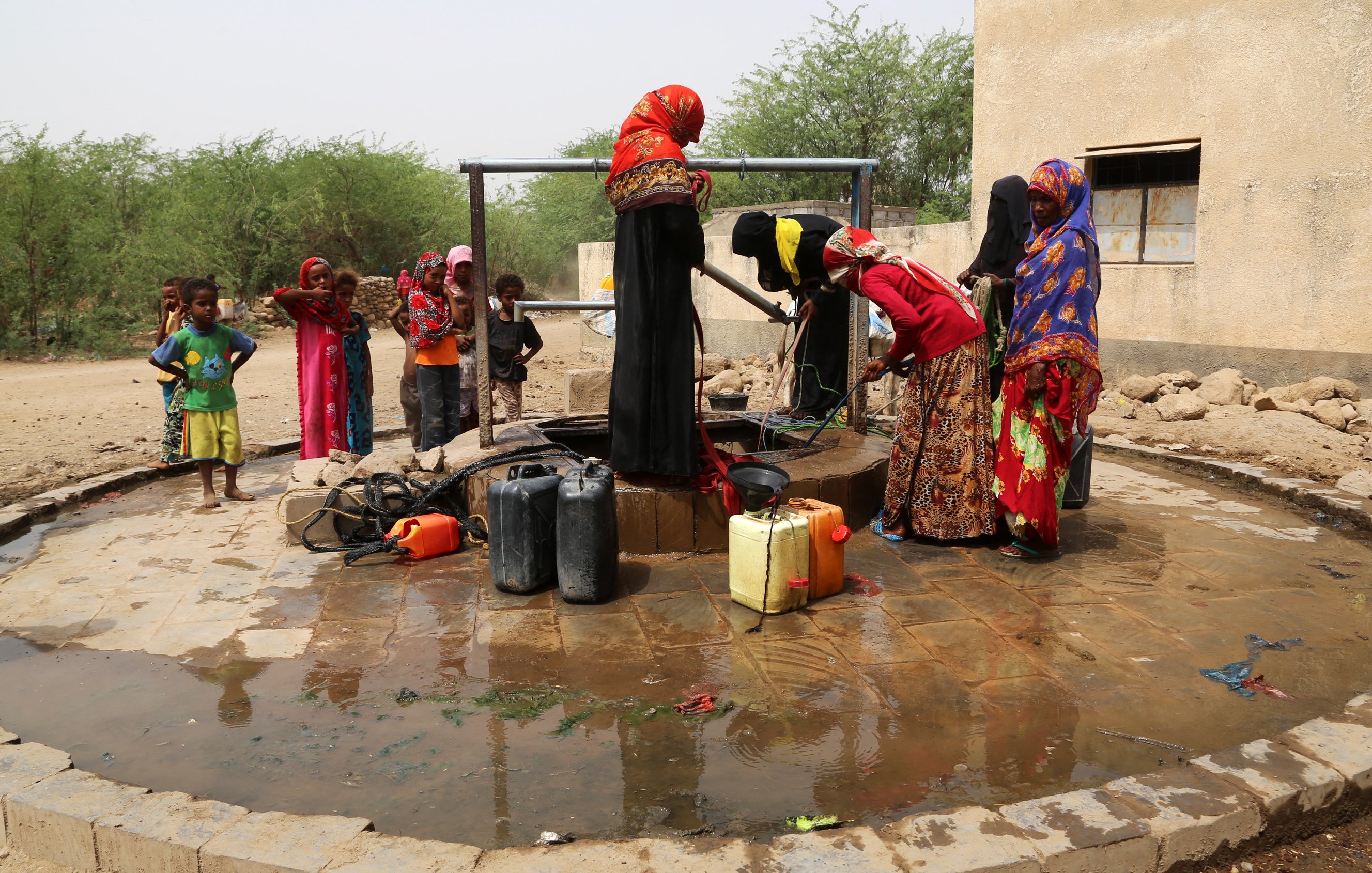 L'image montre un groupe de femmes près d'un puits. Elles sont vêtues de robes colorées et semblent être en train de remplir des conteneurs d'eau. Des enfants jouent autour d'elles. Le sol est humide, et le cadre semble rural, avec des buissons et une structure en arrière-plan. L'atmosphère semble active et communautaire, illustrant l'importance de l'accès à l'eau dans la vie quotidienne.