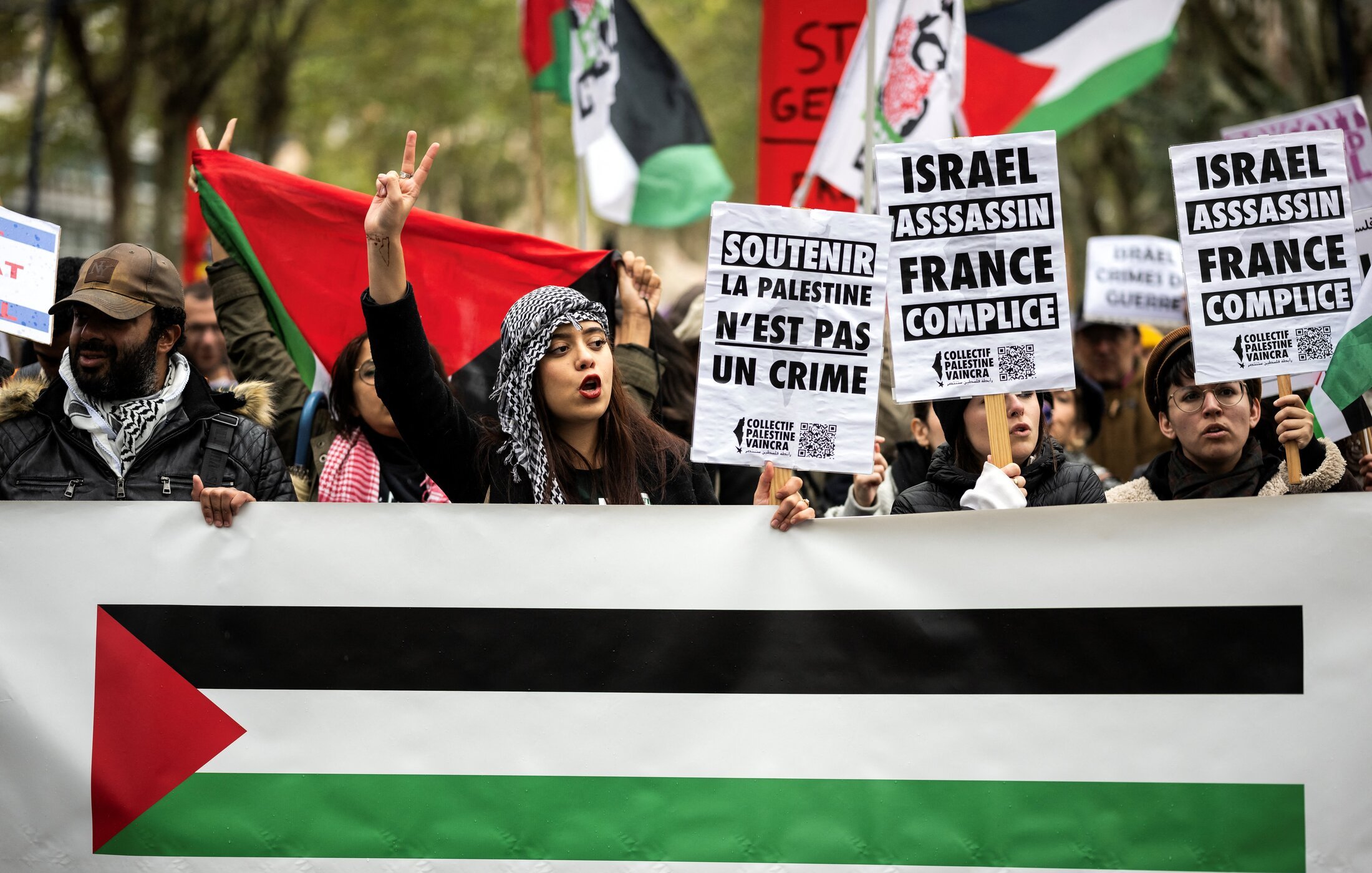 L'image montre une manifestation avec des personnes brandissant des pancartes et des drapeaux. Au premier plan, on voit une femme portant une écharpe avec un motif traditionnel qui exprime un message pacifiste en levant les deux doigts. Les pancartes contiennent des slogans concernant la Palestine, ainsi que des accusations contre Israël. À l'arrière-plan, des drapeaux aux couleurs du drapeau palestinien sont visibles, créant une atmosphère de revendication politique et de solidarité.