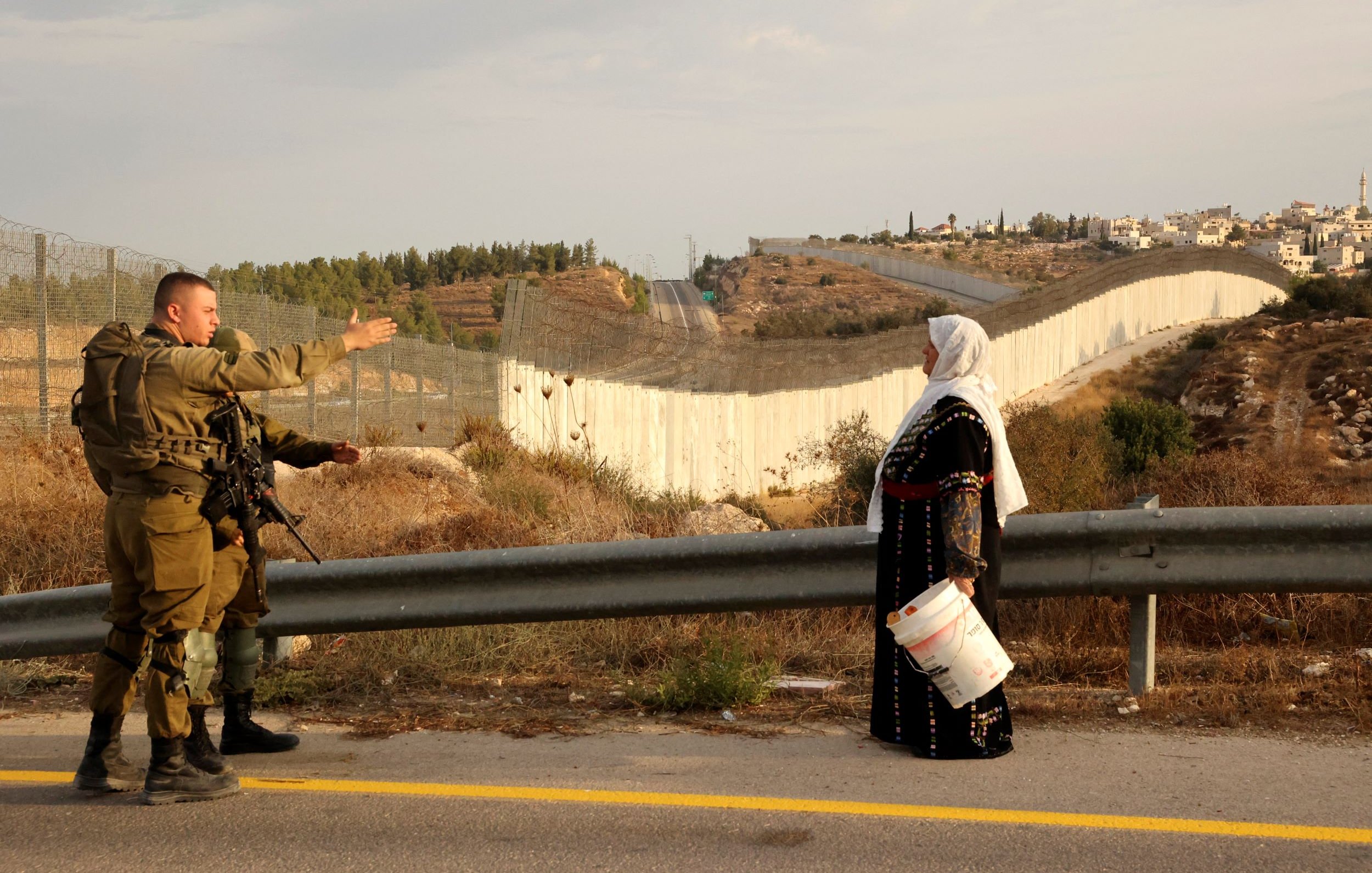 L'image montre un soldat en uniforme qui semble communiquer avec une femme vêtue de manière traditionnelle, tenant un seau. En arrière-plan, on aperçoit un mur ou une barrière qui sépare les deux côtés, avec des collines et des bâtiments au loin. L'environnement semble rural, avec de la végétation et une route. Cette scène évoque un contexte de conflit ou de tension dans une région marquée par des divisions.