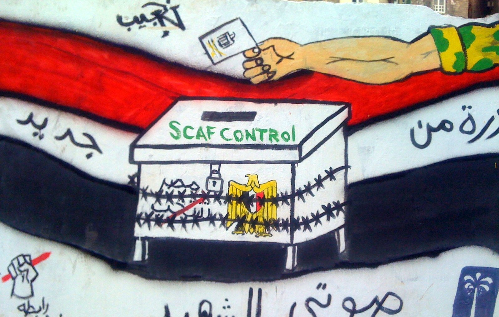 L'image montre une fresque murale avec une boîte de vote entourée de barbelés. Au-dessus de la boîte, on peut voir la phrase "SCAF CONTROL" écrite en vert. À gauche, il y a un poing levé, symbolisant la résistance ou la lutte, et à droite, on peut apercevoir des symboles qui représentent probablement des idéaux de liberté et de changement. La fresque utilise des couleurs vives comme le noir, le rouge et le blanc, qui sont souvent associés à des mouvements politiques. Le message semble critiquer ou commenter le contrôle militaire sur le processus électoral.