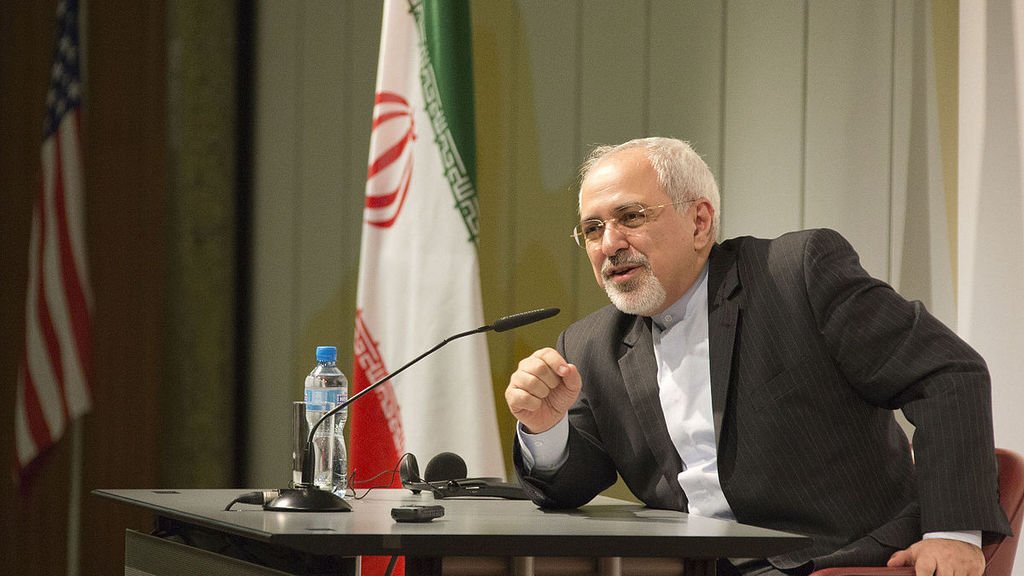 L'image montre un homme assis à une table de conférence, parlant avec animation. Il est habillé en costume, avec une chemise claire. À l'arrière-plan, on peut voir des drapeaux, y compris celui de l'Iran et celui des États-Unis. L'homme semble s'adresser à un public, probablement en répondant à des questions ou en faisant une déclaration importante.